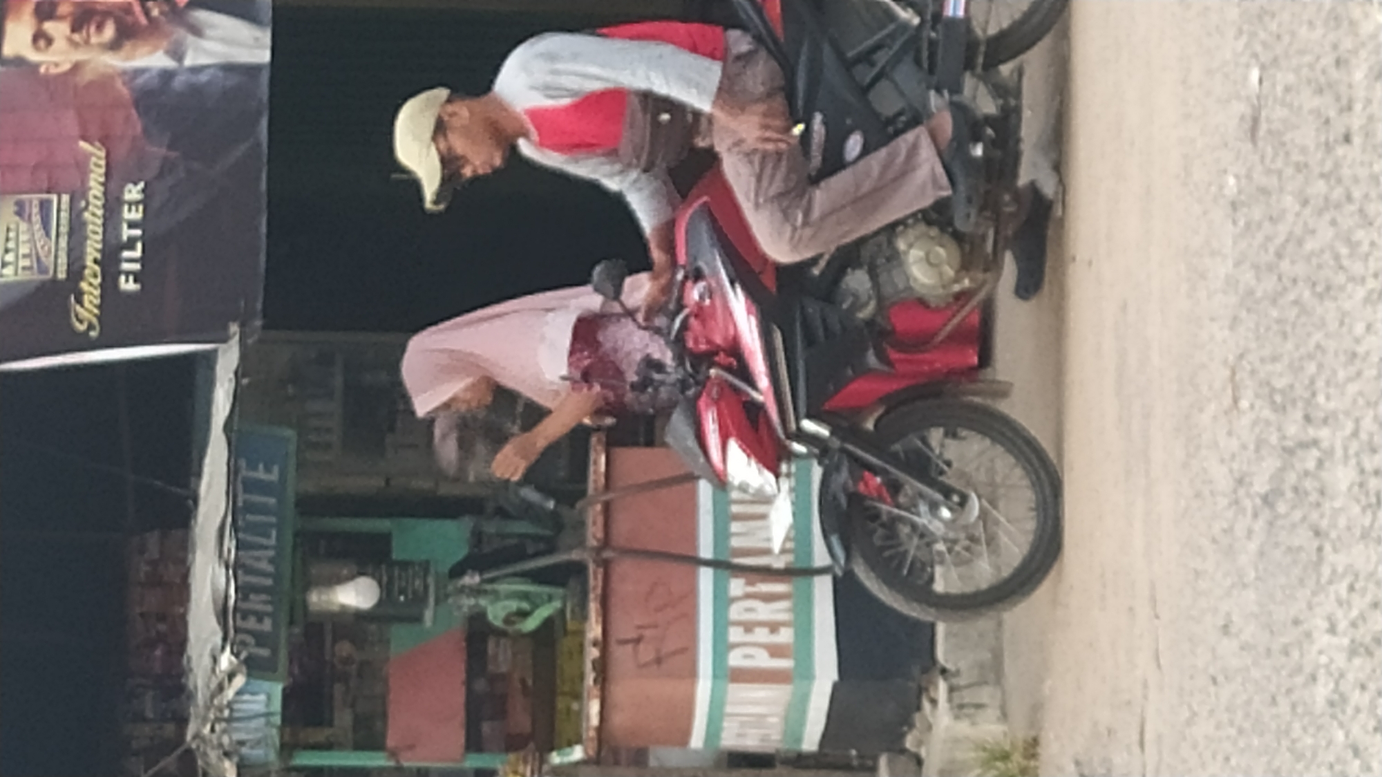 The image captures a street scene in Klapanunggal, Indonesia, likely near a roadside stall. In the foreground, a red motorcycle is parked on a dirt road. A man wearing a light brown baseball cap, sunglasses, a red and gray long-sleeved shirt, and brown pants is seated on the motorcycle, facing left. He has a dark brown fanny pack slung across his chest. To his left, standing behind the motorcycle, is a woman wearing a pink hijab and a patterned long-sleeved top. She is looking down and appears to be interacting with something held in her right hand. Behind them, a small stall is visible, with a sign that reads "PERTALITE" in blue lettering. The stall has a green structure and a red-topped counter. Above the stall, a large banner advertising "International FILTER" with a man's portrait is displayed. The overall environment suggests a casual, outdoor setting in a commercial or semi-urban area.