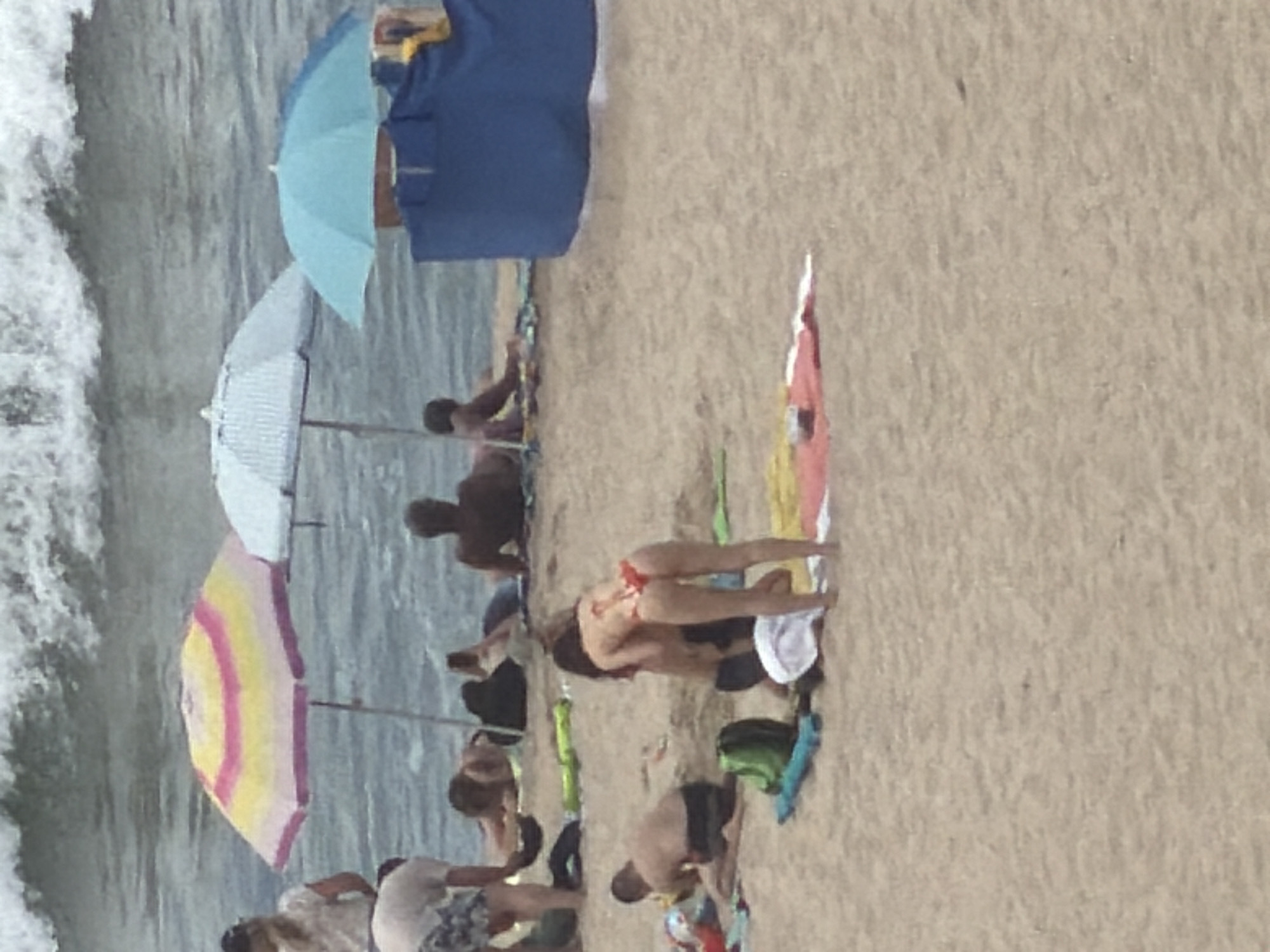 The image shows a crowded beach scene with numerous people and beach umbrellas. In the foreground, a woman in a red bikini is bent over. To her left, a man in dark swim trunks is seated on the sand. Several beach towels and bags are scattered around the foreground and middle ground. In the middle ground, a group of people are lying on the sand, some of whom are under beach umbrellas. The ocean with waves is visible in the background. The location appears to be a public beach.
