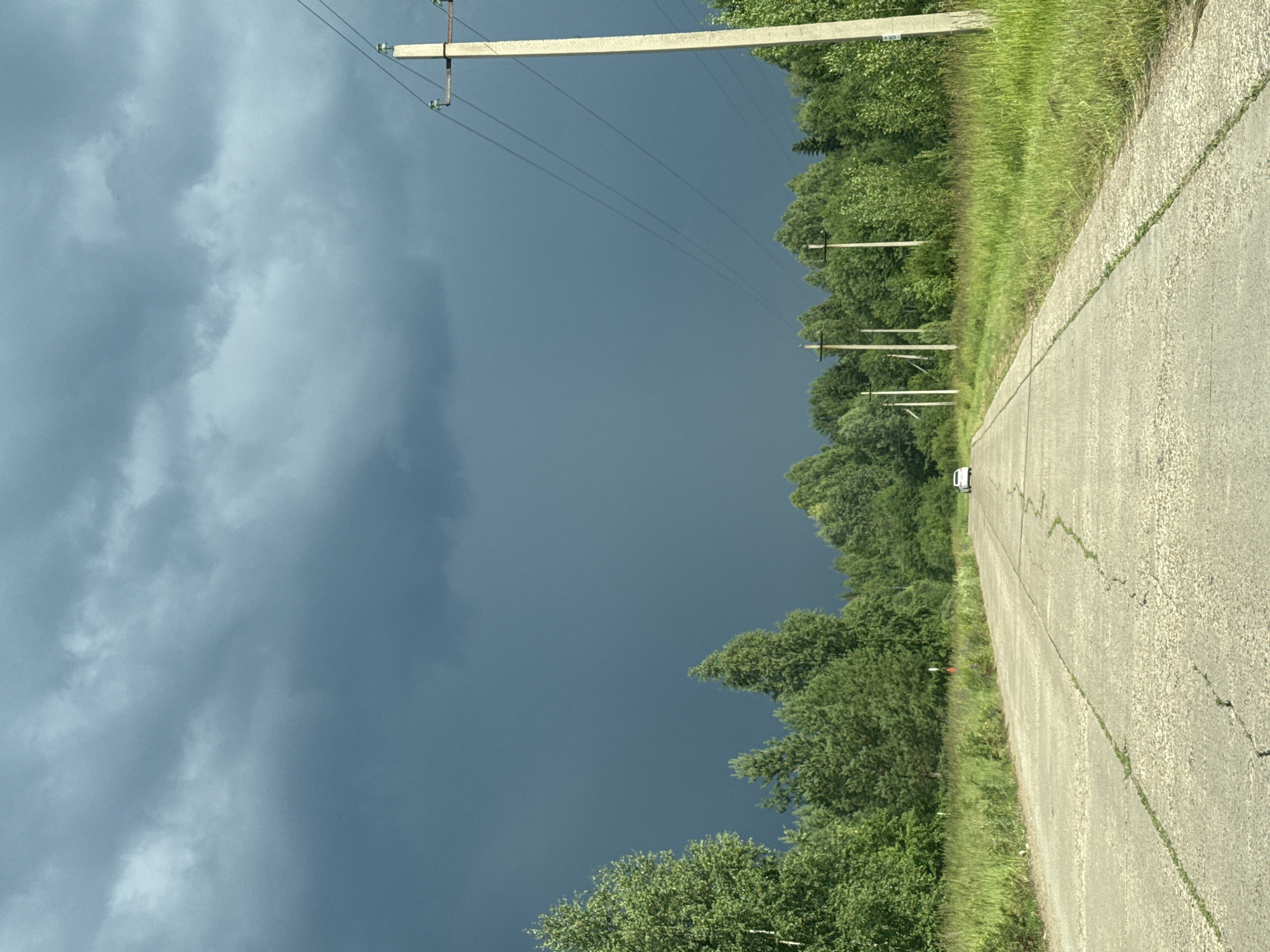 A rural asphalt road stretches into the distance, flanked by a dense forest on either side. The sky above is filled with dark, ominous storm clouds, suggesting an impending downpour. A single white car is visible on the road, moving away from the viewer. Several utility poles with power lines run along the right side of the road. The road surface appears worn, with cracks and patches visible. The overall environment is natural and somewhat desolate, with no signs of human habitation other than the road and power lines. This scene could be located on the outskirts of Chaikovsky, Russia, where such undeveloped landscapes are common.