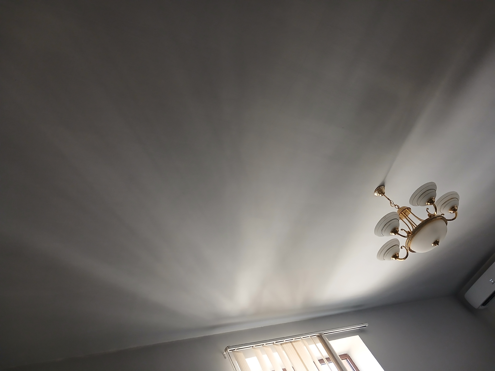 The image depicts an indoor scene, likely a room with a high ceiling. A gold chandelier with five white glass shades is prominently featured, suspended from the ceiling. To the left, a window with horizontal blinds is visible, allowing light to stream in and cast a pattern of light and shadow on the wall. An air conditioning unit is mounted near the ceiling in the lower left corner of the frame. The overall impression is one of a well-lit, possibly modern interior space. There are no people or animals present in the image. The location of Kasakh, Armenia, is not discernible from the visual elements within the photograph.