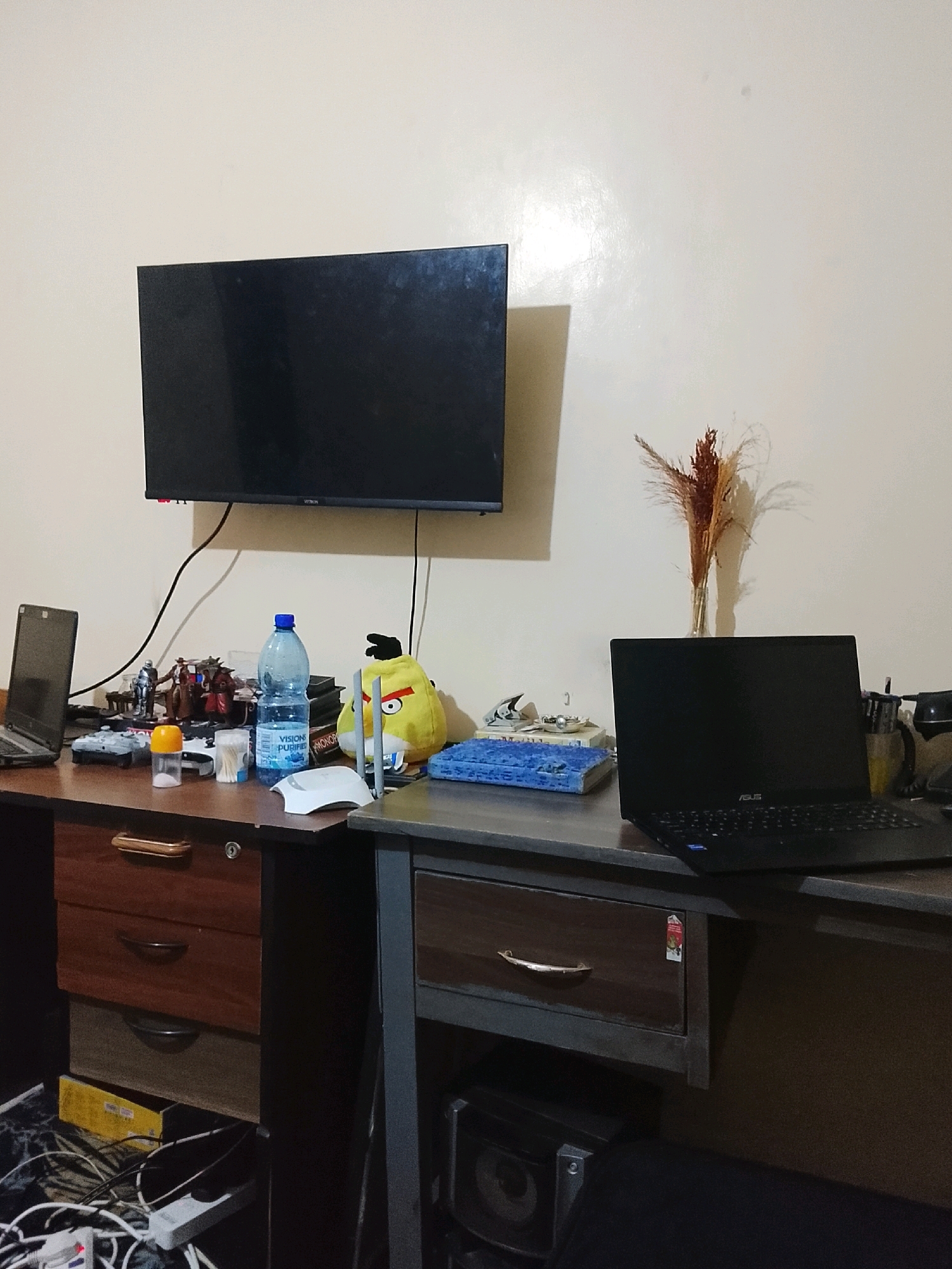 The image depicts a cluttered desk area in what appears to be a residential room, likely in Ongata Rongai ward, Kenya. On the left side of the desk, a black laptop is open, and to its right, a bottle of purified water stands next to some action figures and a yellow plush toy. A flat-screen television is mounted on the wall above the desk. To the right of the desk, another black laptop is open, and a vase containing dried flowers is placed behind it. The desk is made of wood with visible drawers. Various cables and power strips are visible underneath the desk. The overall environment suggests a workspace or study area.