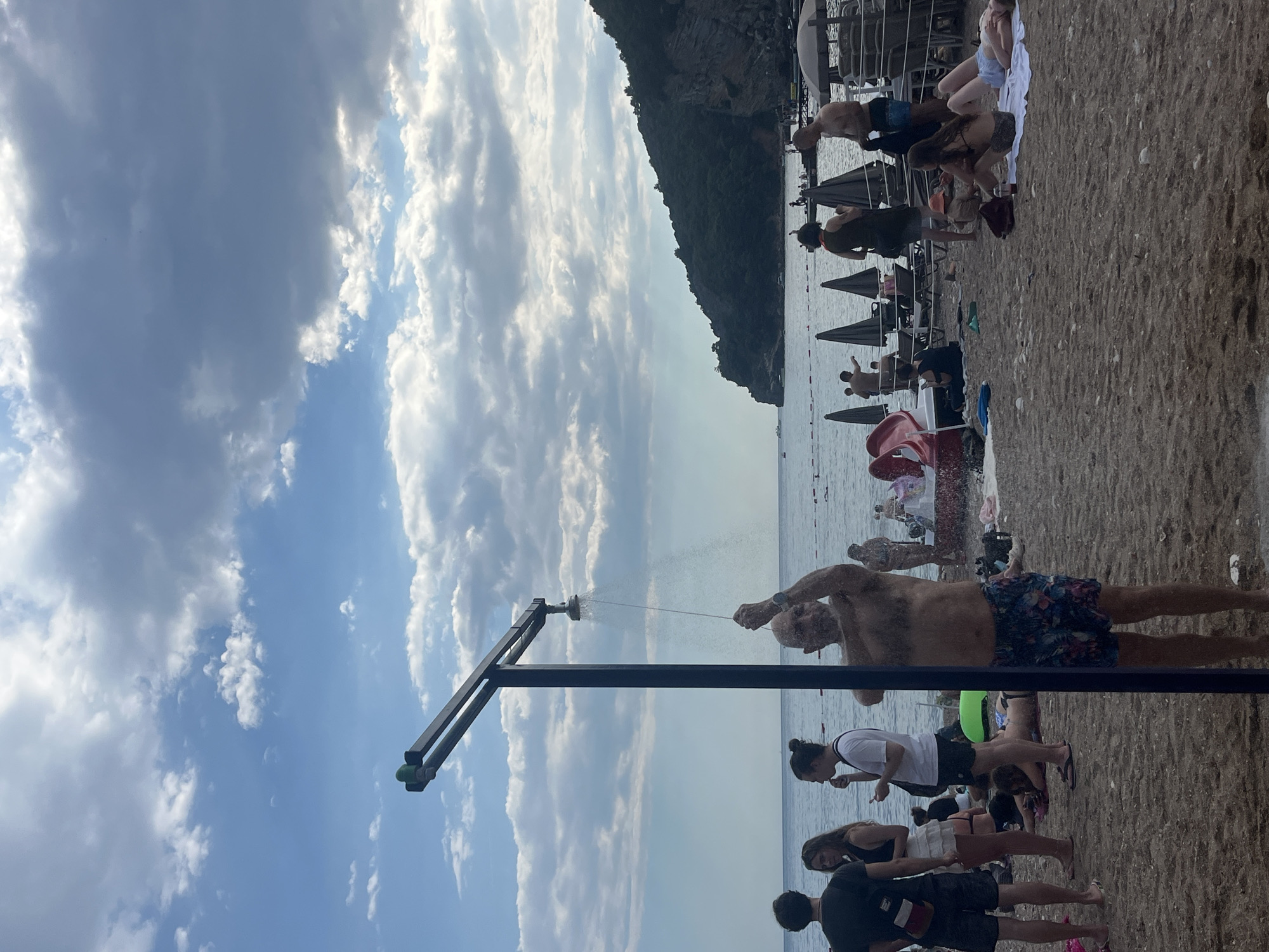 The image depicts a crowded beach scene on a cloudy day, likely in the city of Budva, Montenegro. In the foreground, a man is shown showering outdoors under a metal shower fixture. He is wearing patterned swim trunks and has his hands raised, possibly to wash his hair. Several people are visible on the sandy beach behind him. To his left, three individuals stand together, two women and one man. One woman wears a white top and skirt, while the other wears a dark top and a white skirt. The man on the far left is wearing a black t-shirt and shorts. Further back, other beachgoers are scattered across the sand, some lying down on towels, others standing or walking. Beach umbrellas and lounge chairs are set up in sections of the beach. The sea is calm and extends to the horizon, where a dark, tree-covered hillside dominates the background. The sky is filled with a mix of dark and light clouds, suggesting an overcast or partially cloudy day. The overall atmosphere is one of a typical busy summer day at a coastal resort.