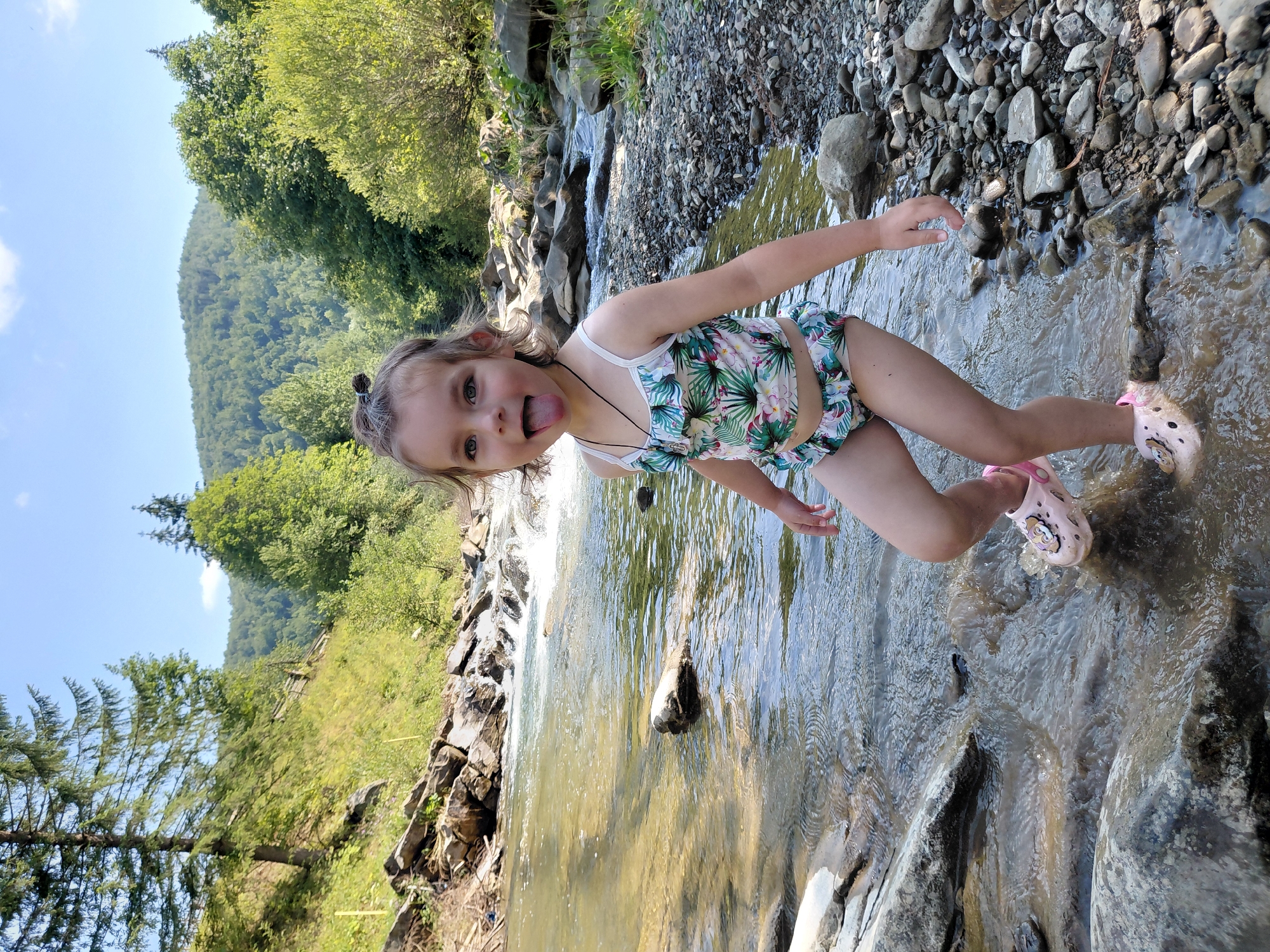 A young girl with blonde hair tied in two buns, wearing a floral swimsuit and pink clogs, stands in a shallow, rocky stream. She has her tongue sticking out, looking playfully at the camera. Her right arm is outstretched towards the water, while her left arm is bent at the elbow. The stream flows over rocks and pebbles, with a small waterfall visible in the background. The surrounding area is lush with green trees and a verdant hillside, suggesting a natural, outdoor setting. The sky is clear and blue, indicating fair weather. The image appears to have been taken during the daytime. The location is identified as Chernyk, Ukraine. There is no visible text in the image.