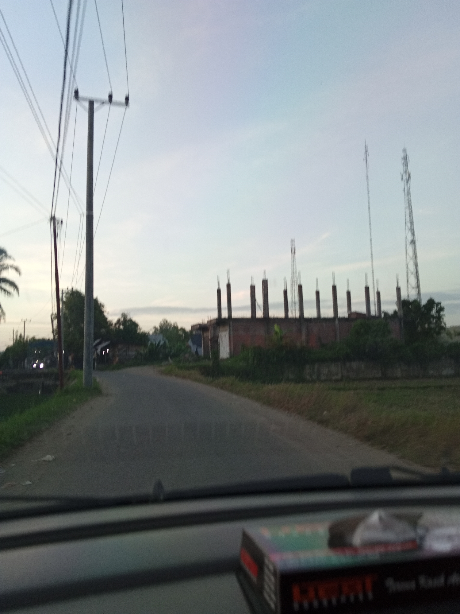 The image depicts a road in what appears to be a rural area, likely located in or near Tingkeummanyang, Indonesia, given the prompt. The perspective is from inside a vehicle looking forward onto the road. The road is asphalt and stretches into the distance, curving slightly. On the left side of the road, there's a grassy verge with utility poles carrying power lines running parallel to the road. On the right, there's a building under construction with exposed concrete columns extending above a brick wall. Beyond the building, two communications towers are visible against the sky. The sky is overcast, suggesting either early morning or late afternoon light. In the foreground, the vehicle's dashboard is visible, with an object (likely a box or container) on it. No people are clearly visible in the scene. The overall impression is one of a developing rural landscape.