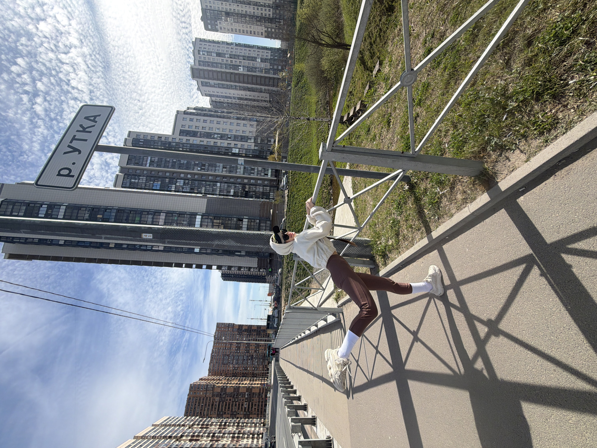 A horizontal eye-level shot depicts a woman in white and brown, leaning on a metal railing on a sidewalk. The shot is oriented so that the sky and tall apartment buildings line the top and bottom edges of the frame. The woman wears a white hooded sweatshirt, brown pants, white sneakers, and white socks. She is arched backwards, holding onto the railing with both hands. The sidewalk is grey and stretches into the distance. To the left, a road sign indicates the area as "P. YTKA," which translates to "Duck River," indicative of the location in Saint Petersburg, Russia. Across the railing, there is grass. In the background, large apartment buildings are visible on both sides of the frame. The sky is blue with cirrus clouds.