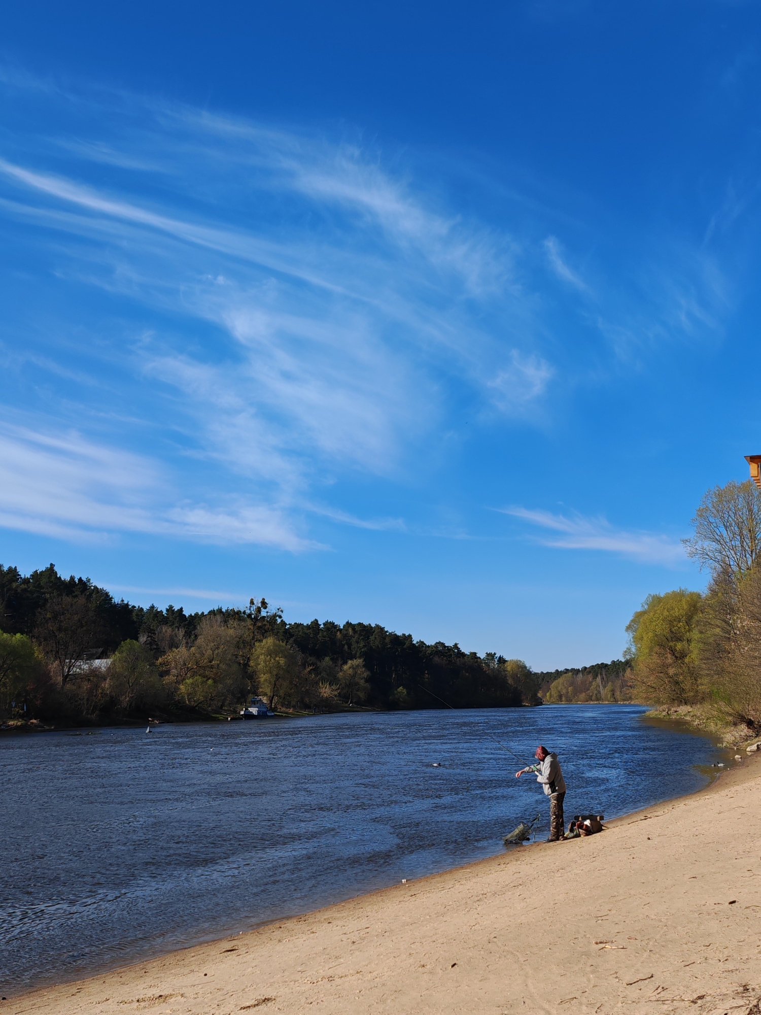A wide-angle outdoor scene depicts a man fishing on the sandy bank of a large river under a clear, bright blue sky with wispy white clouds. The sandy foreground slopes gently down to the dark blue water, which shows subtle ripples and reflections.
A man, wearing a light-colored hoodie, camouflage pants, and a dark cap, stands near the water's edge, holding a fishing rod in a casting or reeling motion. Beside him on the sand are various pieces of fishing equipment, including a green net or bag and a backpack. Several ducks or waterfowl are visible on the water, both near the man and further out.
The opposite bank is densely covered with trees, displaying a mix of early spring green foliage and bare branches. A small blue boat is partially visible, moored near the far bank. In the upper right corner, a glimpse of a reddish-brown structure, possibly part of a building, is seen against the sky.
The weather is bright and sunny, indicative of daytime. The overall impression is one of tranquility and natural beauty, suggesting a recreational activity on a pleasant day. There is no visible text. The setting is a natural landscape, consistent with rural areas like Падлабенскі сельскі Савет, Belarus.