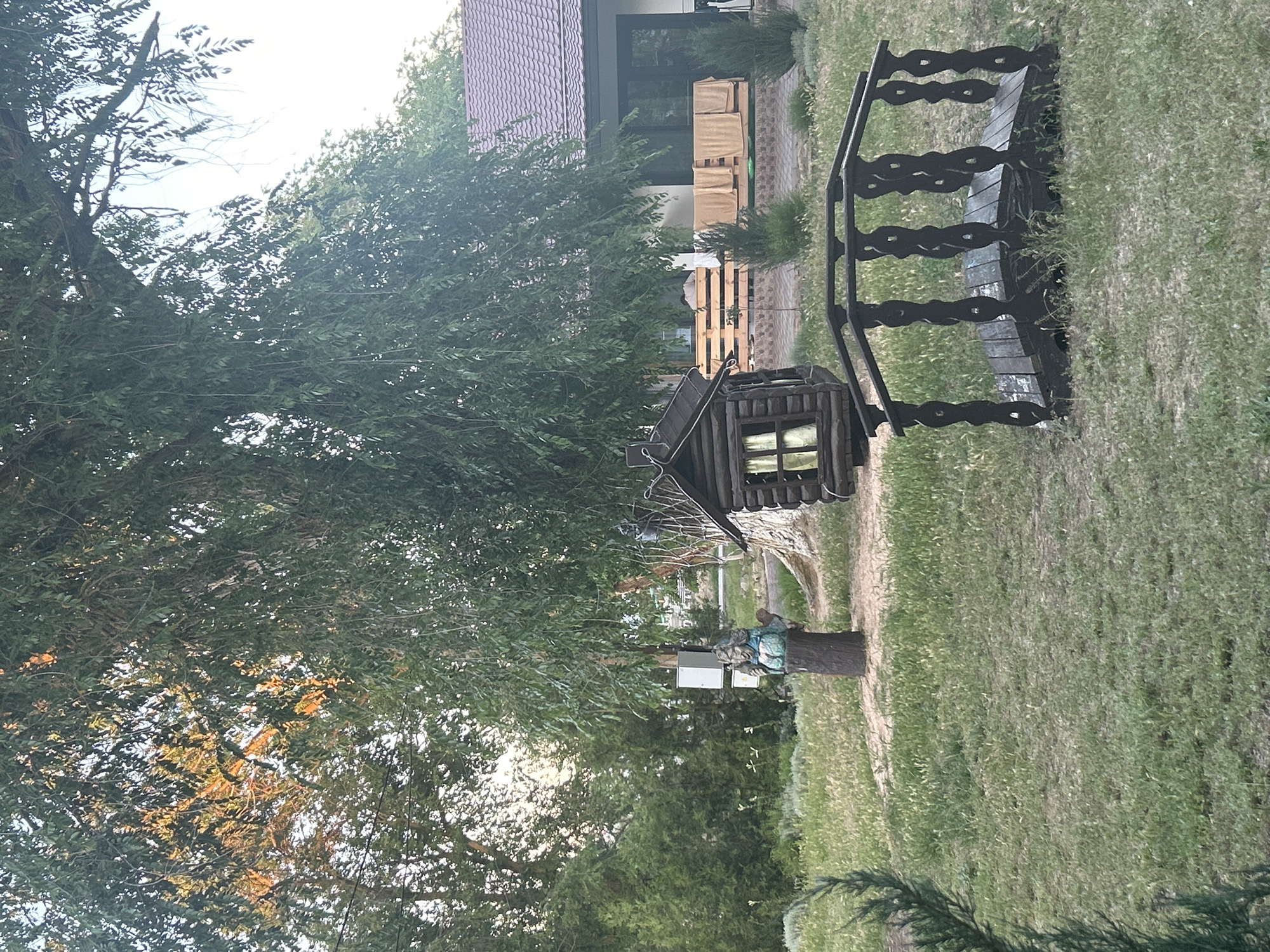 The image depicts a backyard scene in Останинское сельское поселение, Ukraine, featuring a variety of decorative and functional elements. In the foreground, a dark wooden footbridge with intricate railings arches over a patch of grass, suggesting a whimsical garden design. To the left of the bridge and slightly further back, a small, dark wooden birdhouse or decorative shed with a window is visible. A carved wooden figure, possibly a gnome, stands on a tree stump to the left of the birdhouse. In the background, a modern house with a tiled roof and large windows is partially obscured by the branches of a large, leafy tree. Several towels are draped over a wooden railing on the porch of the house, and the surrounding area is covered in grass and various types of bushes and trees, indicating a well-maintained outdoor space. The lighting suggests it is either early morning or late afternoon, with sunlight filtering through the leaves.