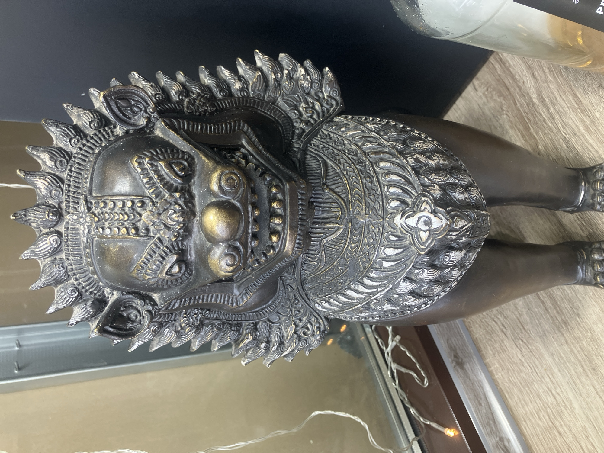 The image shows a bronze-colored, decorative statue of a lion-like creature with ornate armor-like adornments on its head and body. The statue is placed on a light-colored wooden surface near a glass pane and some string lights. A dark object is in the background, possibly a shelf or wall. The location, Chaikovsky, Russia, is extraneous as it cannot be verified from the visual information provided.