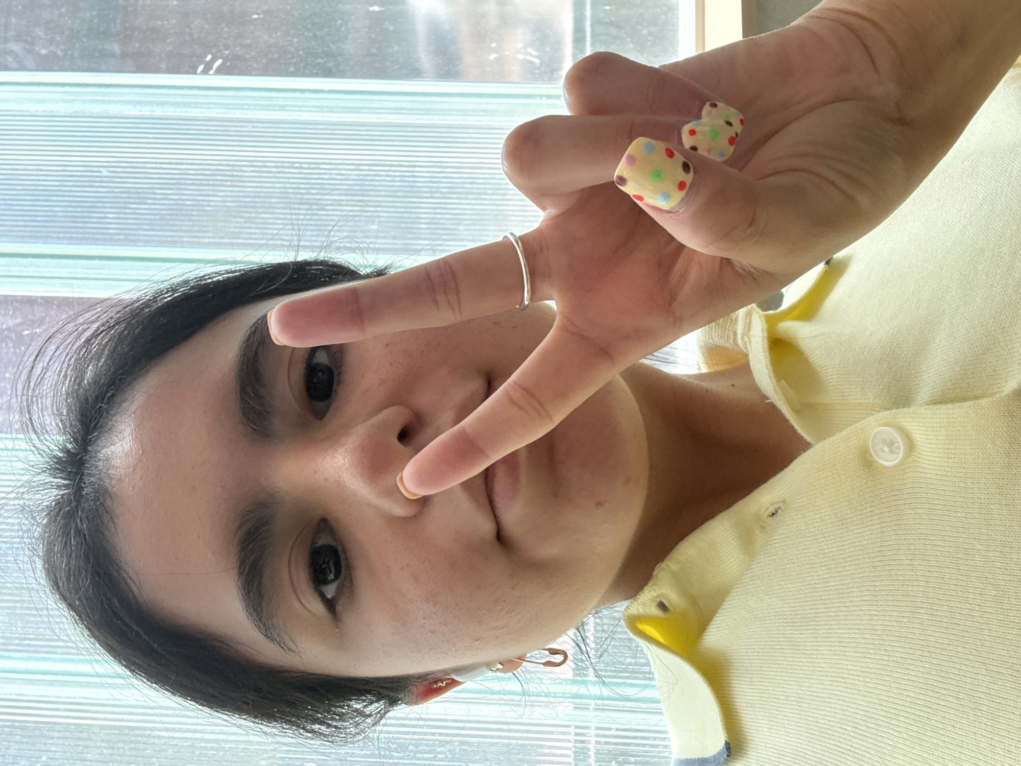 A young woman poses for a selfie, making a peace sign with her fingers. She wears a light yellow cardigan and a thin silver ring on her finger. Her fingernails are painted with a light yellow base and colorful polka dots. She has dark hair and dark eyes. Her left ear is visible and has a safety pin-style earring. The background shows a textured glass window. The location is not identifiable as Austin, United States of America.