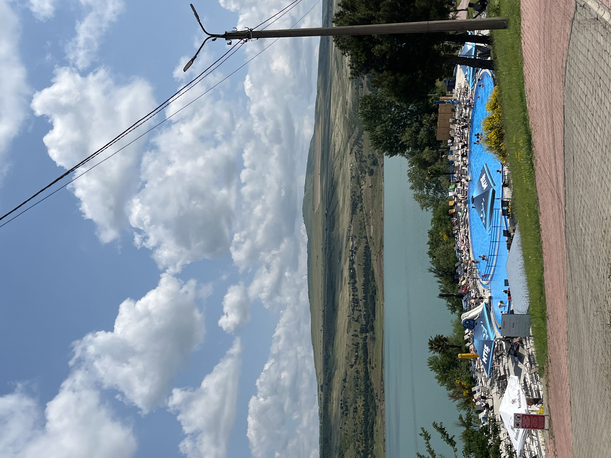 The image depicts a bright, sunny day at an outdoor swimming pool resort located in Tbilisi, Georgia. In the foreground, a paved walkway leads to a spacious pool filled with blue water and several people swimming and lounging. Around the pool are numerous lounge chairs, umbrellas, and some shaded areas. A large blue umbrella with the "MTIS" logo is prominently displayed near a water slide. In the middle ground, a body of turquoise water, likely a reservoir or lake, stretches across the scene. Beyond the water, rolling hills covered in green grass and scattered trees form the landscape, leading to a distant mountain range under a sky filled with white, fluffy clouds. A street lamp and overhead wires are visible in the right side of the frame, suggesting the presence of infrastructure near the resort. The overall impression is one of a recreational area enjoying pleasant weather.