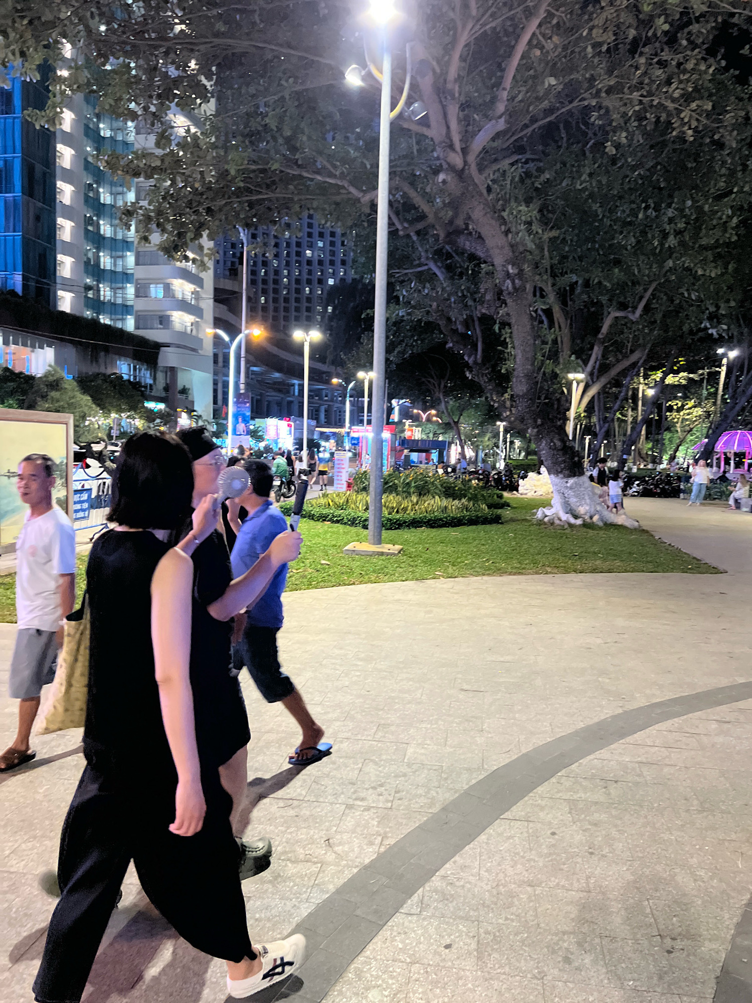 The image depicts a nighttime scene in Nha Trang, Vietnam, likely a public park or promenade. In the foreground, a person wearing a black outfit and white sneakers walks to the left, with their left hand extended. To their right and slightly behind, another person in a black outfit with a black head covering is holding a small portable fan near their face. To the left of the main subjects, a man wearing a white t-shirt and light-colored shorts walks away from the camera.
The environment features a paved walkway with a curved grey band. In the mid-ground, there is a grassy area with several trees, some with their bases whitewashed. In the background, modern buildings with illuminated windows rise up, suggesting an urban setting. Streetlights are visible, illuminating the scene. Other people are present in the mid-ground and background, some walking, and others near parked motorcycles. A purple structure, possibly a gazebo or an awning, is visible in the far right background. The overall atmosphere is that of a lively evening public space.