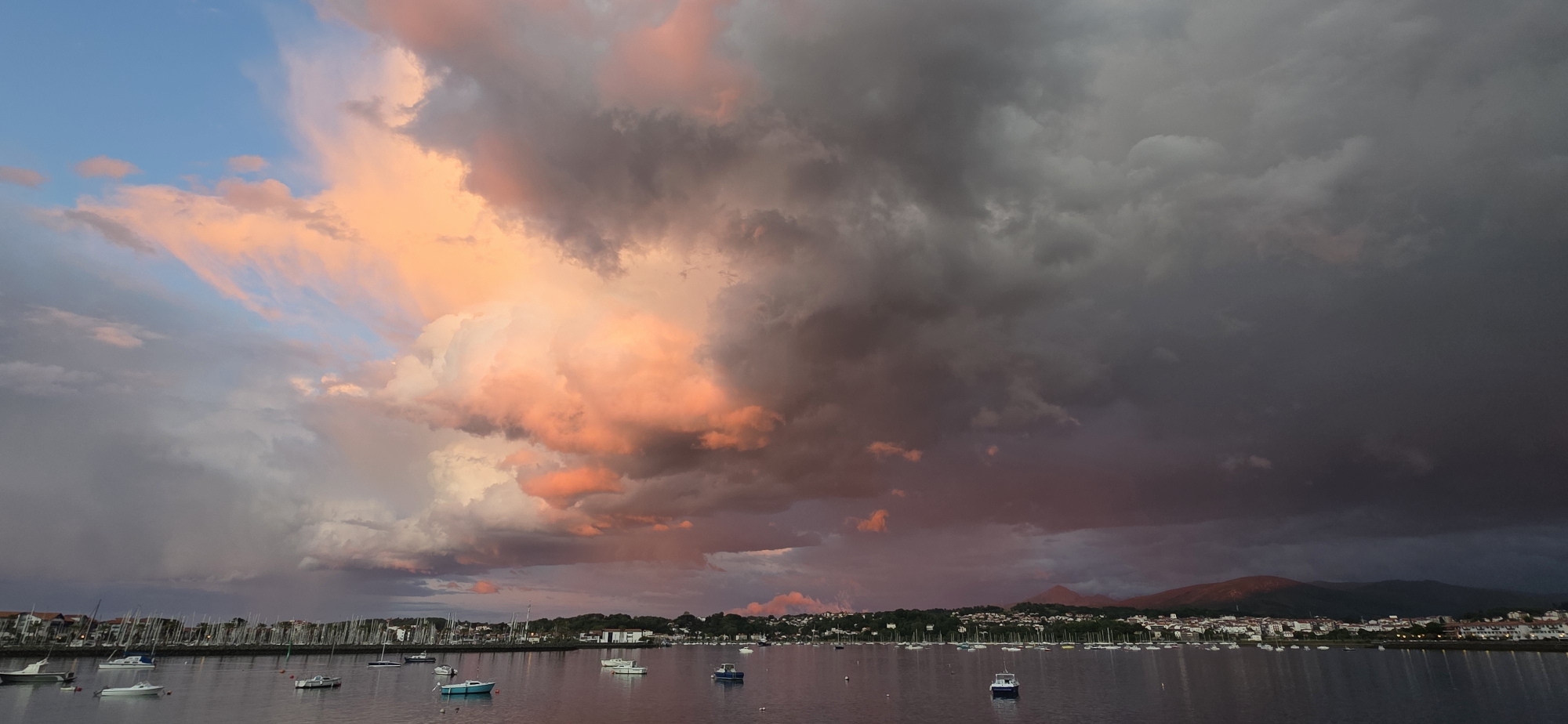 The image captures a panoramic view of Hondarribia, Spain, at dusk. The sky dominates the scene, exhibiting a dramatic contrast between a vibrant sunset on the left side and dark, ominous storm clouds on the right. The setting sun casts a warm, orange-pink hue on the clouds, creating a striking visual effect.
Below the sky, a body of water reflects the colors of the sky and the lights from the town. Numerous sailboats and small boats are scattered across the water, some moored and some adrift. A coastline is visible in the middle ground, featuring buildings, trees, and vegetation, suggesting a town or settlement along the shore. Beyond the town, rolling hills or mountains are visible on the horizon, some tinged with the colors of the sunset. The overall atmosphere is one of tranquility juxtaposed with the power of nature.