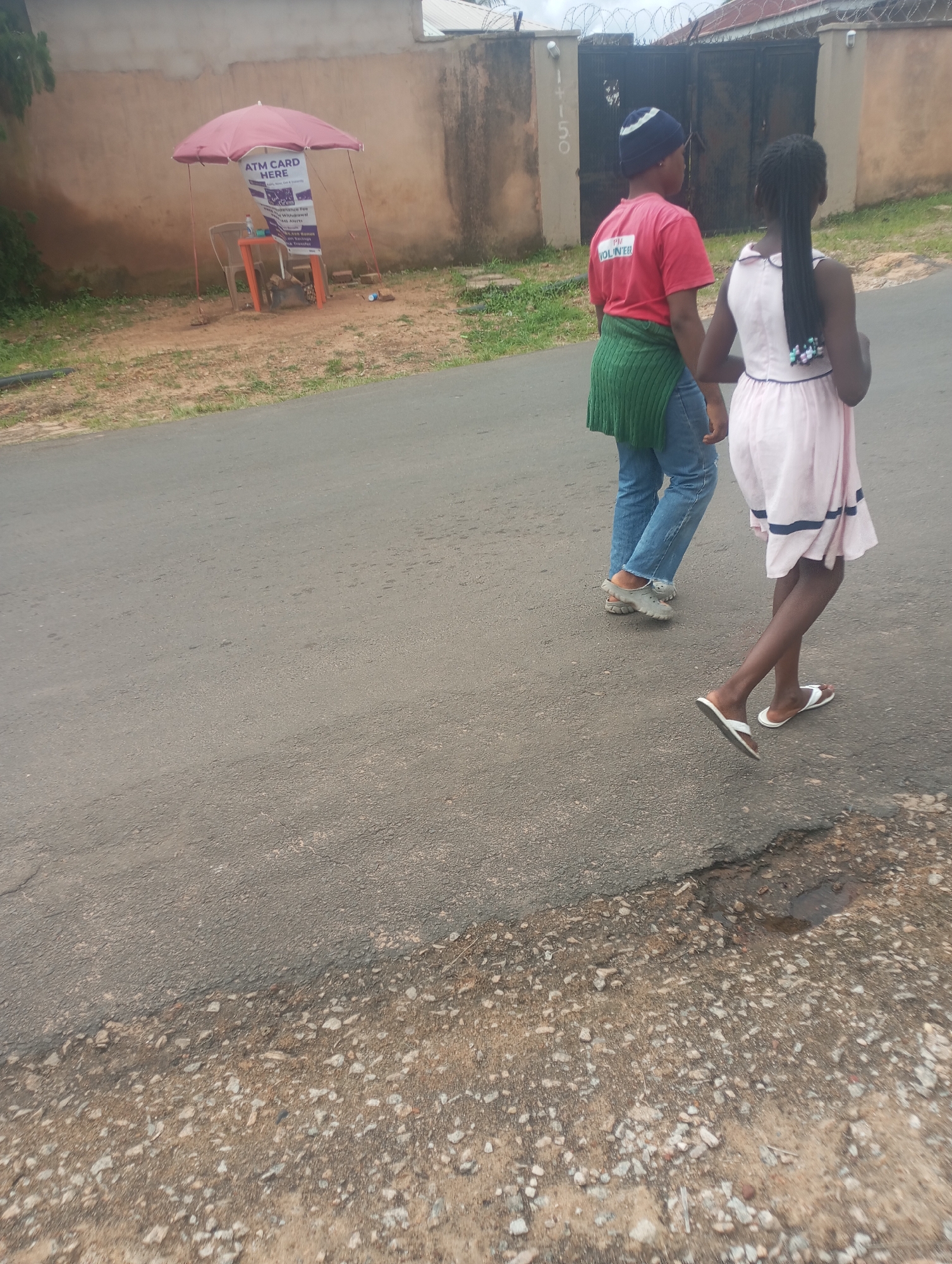 Two young people, a girl and a boy, are walking on a paved road in an urban environment, likely in Jos, Nigeria, based on the visible context. The girl, in the foreground, wears a light pink dress with dark blue trim and white flip-flops. The boy, walking slightly ahead and to the left, is dressed in a red t-shirt with the word "VOLUNTEER" visible on the back, blue jeans, a green sweater draped around his shoulders, and grey clogs. They are walking away from the camera.
To their left, set back from the road, is a small roadside stall. It features a pink umbrella providing shade over a plastic chair and a small orange table. A sign is propped up on the table, advertising "ATM CARD HERE" with details about services offered, suggesting a small business operation. The stall is situated on a patch of unpaved ground with scattered debris, adjacent to a textured wall. In the background, a dark metal gate with barbed wire atop it signifies a boundary or property entrance. The overall environment suggests a typical street scene in a Nigerian city, with a mix of paved roads, unpaved areas, and roadside amenities.