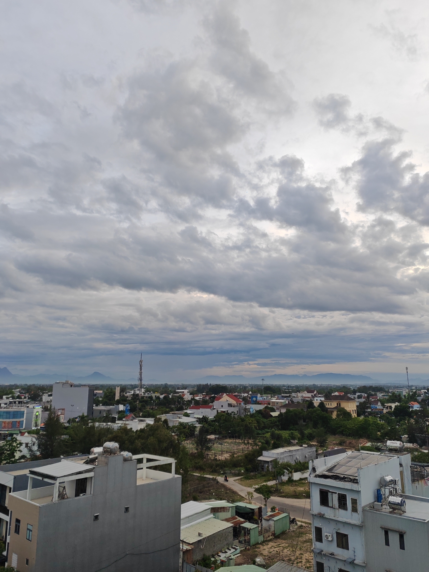 This is an eye-level, medium shot of a city skyline in Đà Nẵng, Vietnam, under a cloudy sky. The foreground is dominated by the tops of several buildings, some of which appear to be residential or commercial structures with multiple stories. One prominent building in the lower right is light blue and has visible rooftop equipment, including water tanks and solar panels. To its left is a gray building with a flat roof and several windows. In the middle ground, there is a mix of buildings, trees, and a cell tower. The buildings in this section are varied in style and color, with some featuring red tile roofs. The background shows a distant mountain range against the hazy sky, with some light breaking through the clouds on the right side of the frame. The overall impression is of a somewhat overcast day in an urban environment with glimpses of natural landscapes.