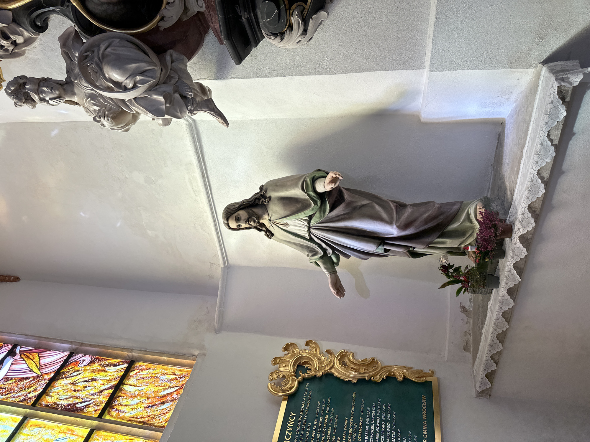 The image depicts an interior shot of what appears to be a church or religious building in Wrocław, Poland. A statue of a male figure, possibly a saint or religious figure, is prominently displayed in a niche with floral arrangements. A plaque with inscriptions is visible below the statue, listing the names of those involved in the renovation of St Elizabeth's Church in Wrocław from 2018-2022. The environment includes elements like stained glass windows and architectural details. Another statue is visible in the top left corner of the image.