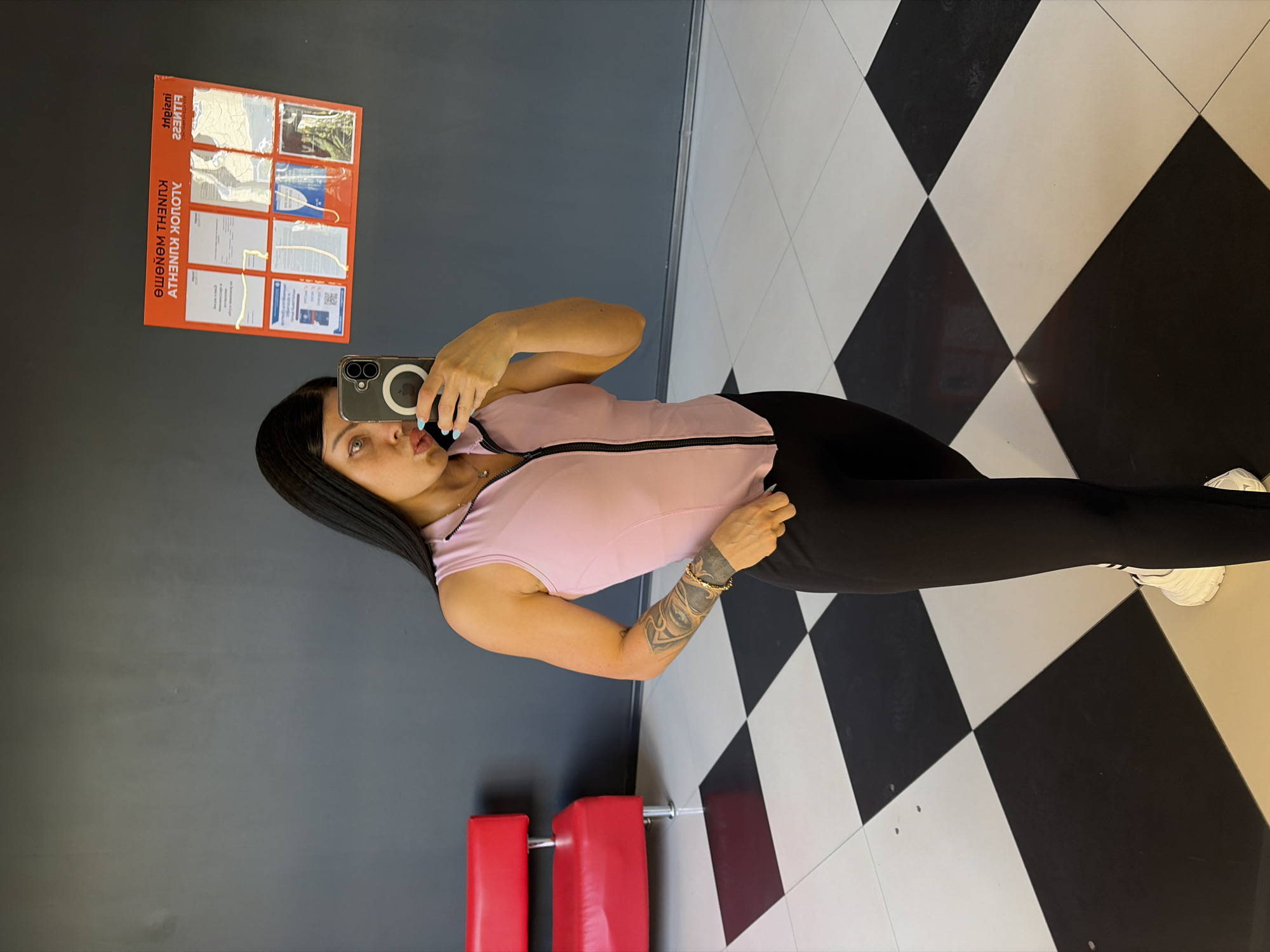 A woman is taking a selfie in a gym. She is wearing a pink sleeveless top with a black zipper and black leggings. She has dark hair and an arm tattoo. The floor is a black and white checkerboard pattern. In the background, there is a grey wall with a poster showing various images and text, which appears to be in Cyrillic. Two red gym equipment pieces are visible in the lower left corner. The lighting suggests it could be daytime. The woman's expression is playful as she pouts for the camera. The poster on the wall contains text that reads, "ЛОШОК КУНЕЧНДУ КИЧКНЛ МЭНЭМЭ".