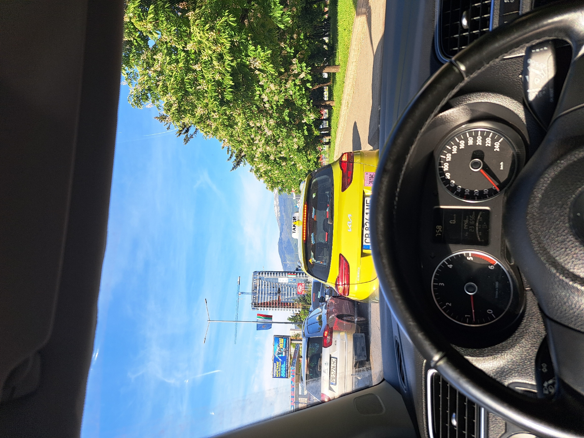 The image, taken from inside a car, shows a traffic jam in Sofia, Bulgaria. A yellow taxi is directly in front, displaying license plate "CB 8261 ME". The car's dashboard is visible, showing the speedometer reading 0 km/h. Outside, a clear blue sky is present, with modern high-rise buildings in the distance, indicating an urban area. A Bulgarian flag is visible. The car is stopped on a road lined with trees on one side. A billboard is visible on the left side of the road. A red car and a white Peugeot car are also visible in the traffic line. The car is a Volkswagen brand based on the visible steering wheel and dashboard elements.