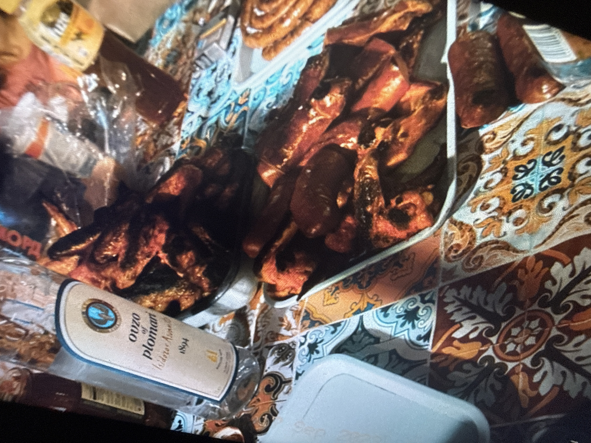This is a close-up, high-angle, medium-shot photograph taken at a picnic table, possibly in Sofia, Bulgaria. The scene is set outdoors, with a vibrant, patterned tablecloth in shades of orange, blue, and white covering the table. The photograph focuses on a spread of food and drinks, suggesting a meal is in progress or has recently concluded.
Dominating the foreground and mid-ground are various food items, primarily sausages and what appear to be pieces of grilled or fried meat. These are arranged on white platters and directly on the tablecloth. The sausages are a deep reddish-brown, some appearing plump and others slightly shriveled. The meat pieces are dark brown and irregularly shaped, with some showing charring. A white plastic container is visible in the lower left quadrant, partially obscuring the tablecloth.
To the left of the food, a clear glass bottle of ouzo is prominently displayed. The label is light beige with blue and white text and features a circular emblem with a blue depiction of a bird or winged creature. The text on the label reads "ouzo of plomari" and "Isidro Arrivas," with the year "1894" also visible. The bottle appears to contain a clear or very lightly colored liquid.
In the background, other items are visible, including a plastic-wrapped package, possibly containing more food, and a yellow bottle with a dark liquid, potentially a sauce or another beverage. The overall impression is one of a casual outdoor gathering, likely a picnic or barbecue, with an emphasis on savory food and alcoholic beverages. The specific location in Sofia, Bulgaria, is not discernible from the visual elements alone.