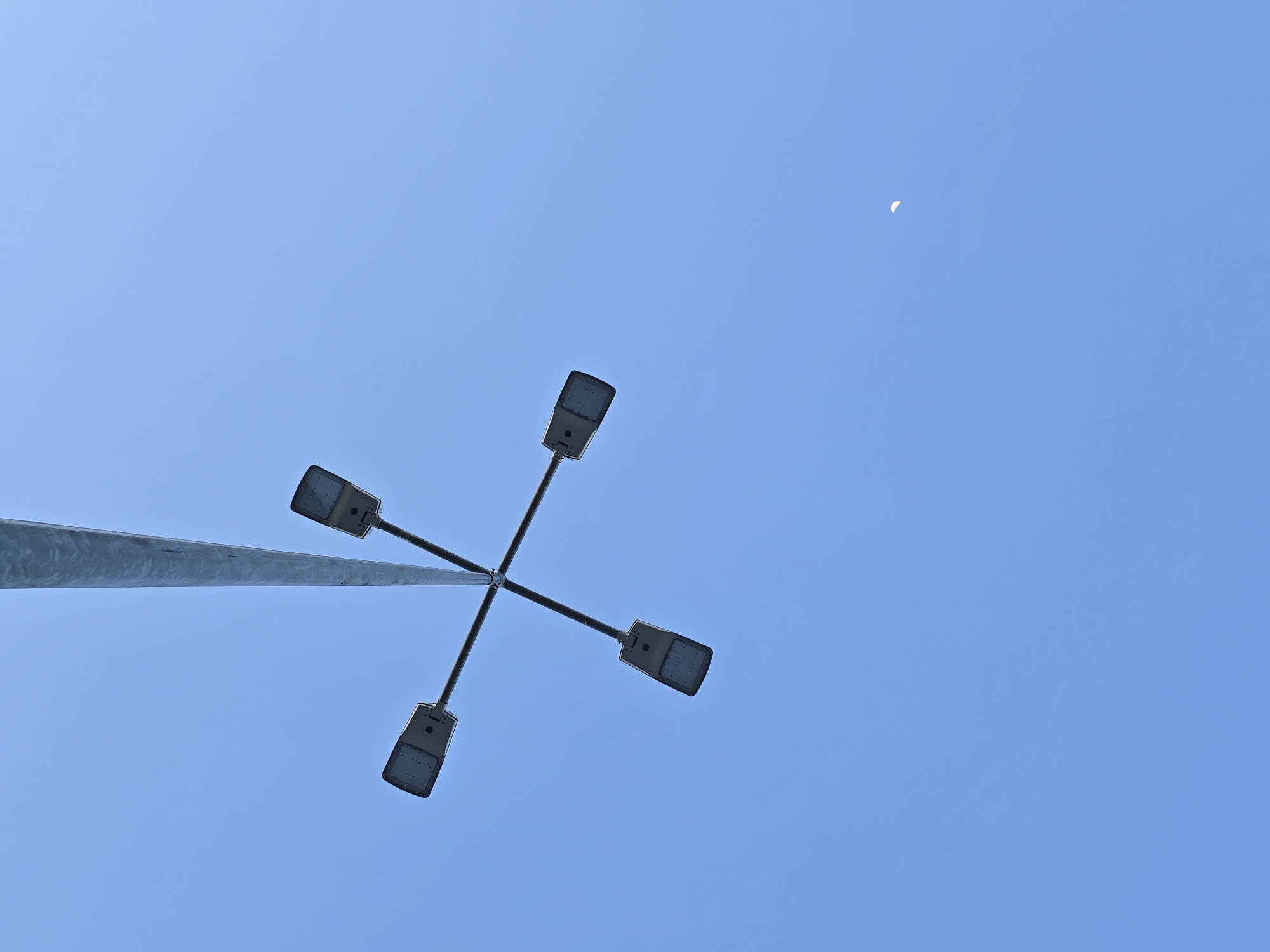 The image captures an upward view of a modern street light against a clear, bright blue sky. A metallic, possibly galvanized, pole extends diagonally from the lower left portion of the frame towards the center. Attached to the top of this pole are four dark-colored, rectangular LED luminaires, also known as street light fixtures. They are arranged in a cross-like configuration, pointing outwards in different directions. Each luminaire features a grid-like pattern on its surface, indicating multiple LED bulbs. In the upper right quadrant of the vast blue sky, a small, faint white crescent moon is distinctly visible. The overall brightness and color of the sky suggest it is daytime, likely early morning or late afternoon. The weather appears clear with no clouds. No people or visible text are present in the frame.