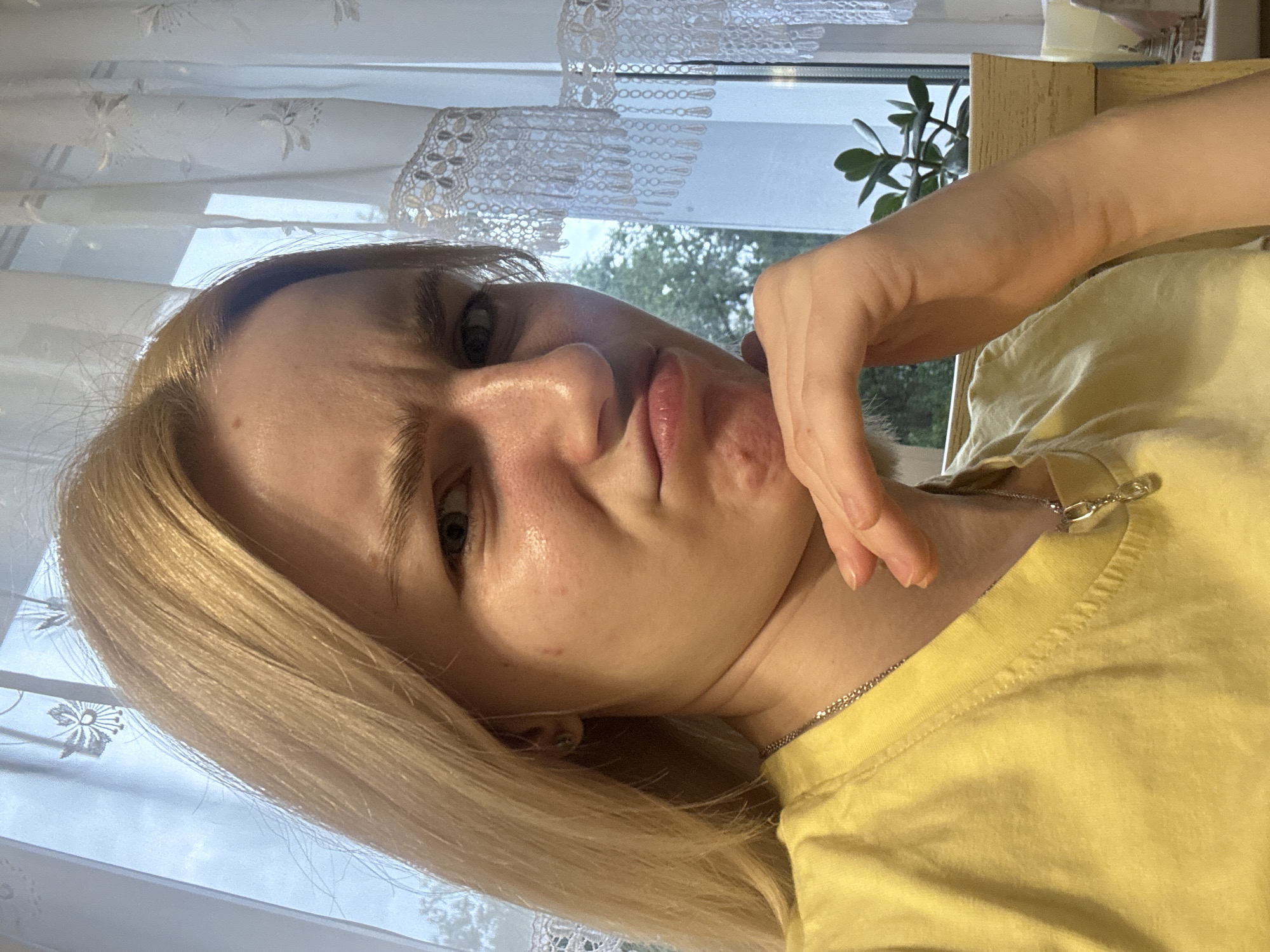 A blonde-haired woman with a yellow shirt is looking at the camera. She is resting her chin on her hand. She has an annoyed expression on her face. The background is blurry but appears to be a room with a window and curtains. The woman is located in Sinyavino, Russia.