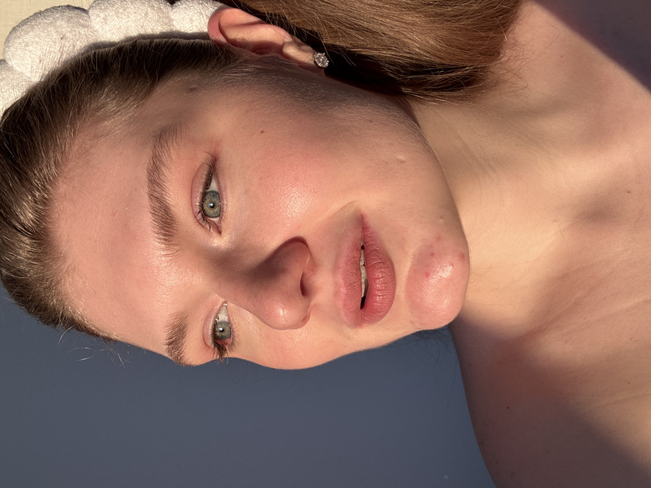 A close-up portrait depicts a fair-skinned female individual, likely a young adult, situated in Kazan, Russia. Her light brown hair is pulled back, partially revealing a white, scalloped headband. She wears a single clear-stoned stud earring in her visible right ear. Her green-blue eyes are directed towards the left side of the frame. Her lips are slightly parted and appear to have a matte, reddish-brown application. A noticeable blemish is present on her chin. Her bare shoulders are partially visible in the lower section of the frame. Strong, warm, directional light illuminates the right side of her face and body, creating distinct shadows on the left. The background consists of an out-of-focus, plain blue-gray surface.