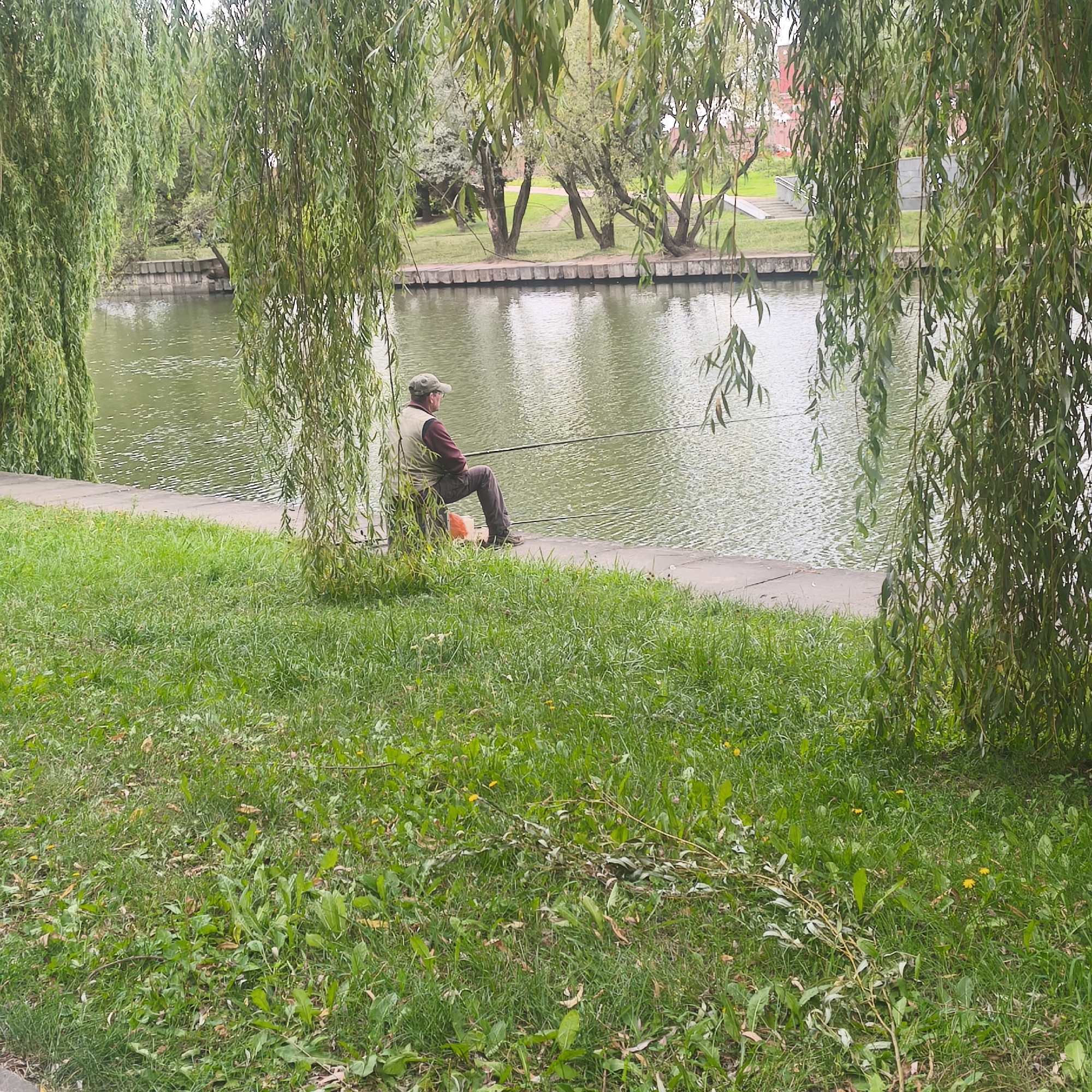 A man is fishing by a river in Minsk, Belarus. He is sitting on a folding chair on a paved bank, with a fishing rod in his hands. The river is calm and reflects the trees on the opposite bank. The foreground is a grassy area with some fallen branches. The overall scene is peaceful and serene.