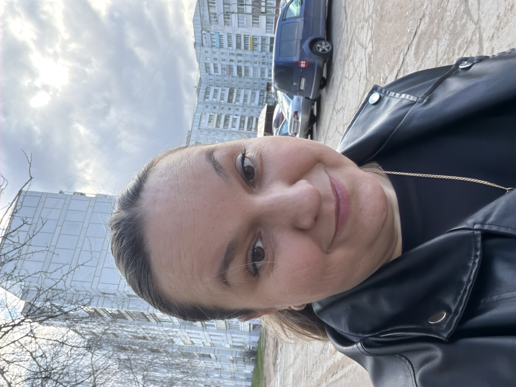 A woman is featured in a self-portrait taken outdoors, smiling subtly and looking directly at the camera. She has dark eyes with visible mascara and is wearing light pink lipstick. Her hair is pulled back, and she is dressed in a black leather jacket over a black top, complemented by a delicate gold chain necklace. The image is framed from a tilted perspective, capturing her face prominently in the foreground.
The background reveals an urban environment, consistent with Chaikovsky, Russia. To the left, a modern building with a distinctive glass facade is partially visible, alongside bare tree branches, suggesting a colder season. To the right, a large, multi-story residential apartment block with numerous windows stands behind a paved area. Several vehicles are parked on the pavement, including a blue van and a silver sedan. The sky above is overcast with a mix of grey clouds and brighter patches where the sun is breaking through. The overall lighting indicates it is daytime. No clearly readable text is visible within the image.