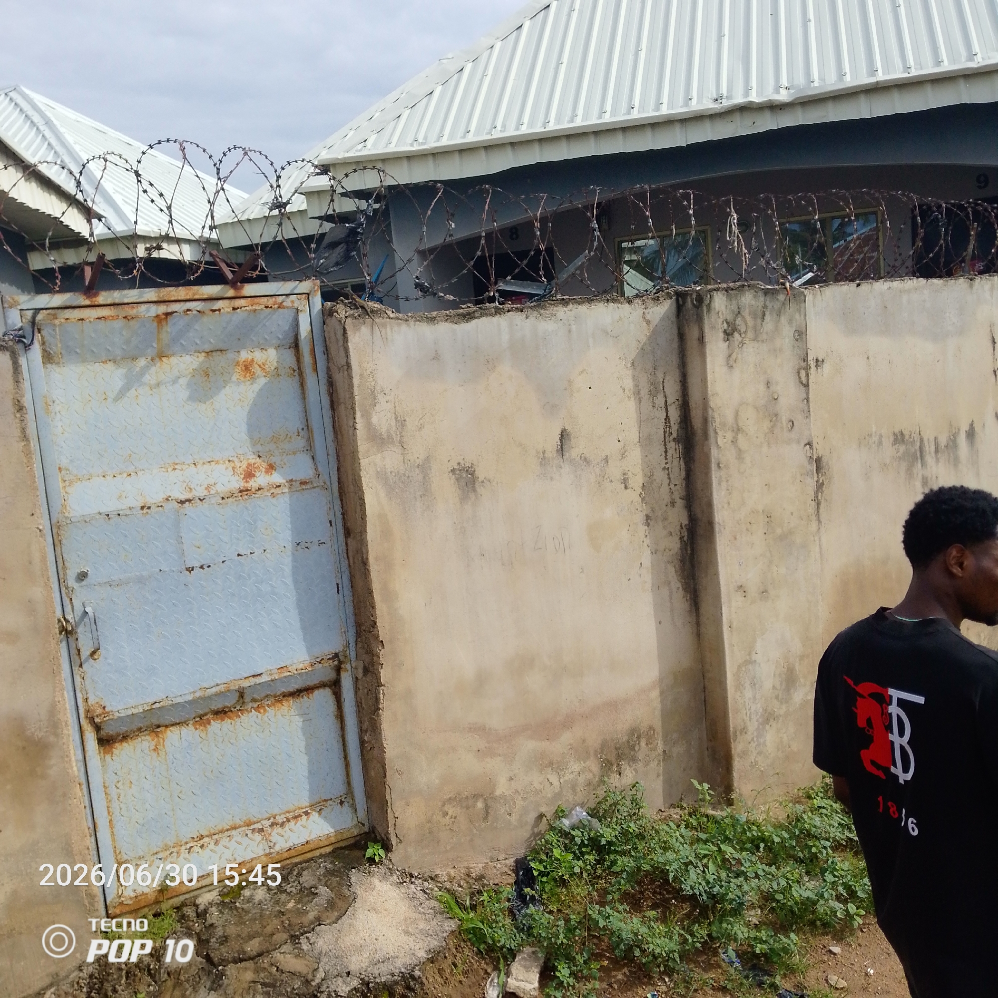 The image shows a man standing in front of a concrete wall topped with barbed wire. The wall is weathered and stained, with some faint writing visible on its surface. Behind the wall, a building with a corrugated metal roof is partially visible. A rusty metal gate is on the left side of the image. Greenery, including small plants and weeds, grows around the base of the wall and the gate. The sky appears cloudy, suggesting an overcast day. The man is wearing a black t-shirt with a red and white graphic design and the number "1816" printed on the back. A timestamp "2026/06/30 15:45" and "TECNO POP 10" are visible in the bottom left corner of the image. The scene appears to be outdoors in a somewhat neglected urban or semi-urban environment. No specific activity or event is clearly taking place; it is a still image of a person and their surroundings.