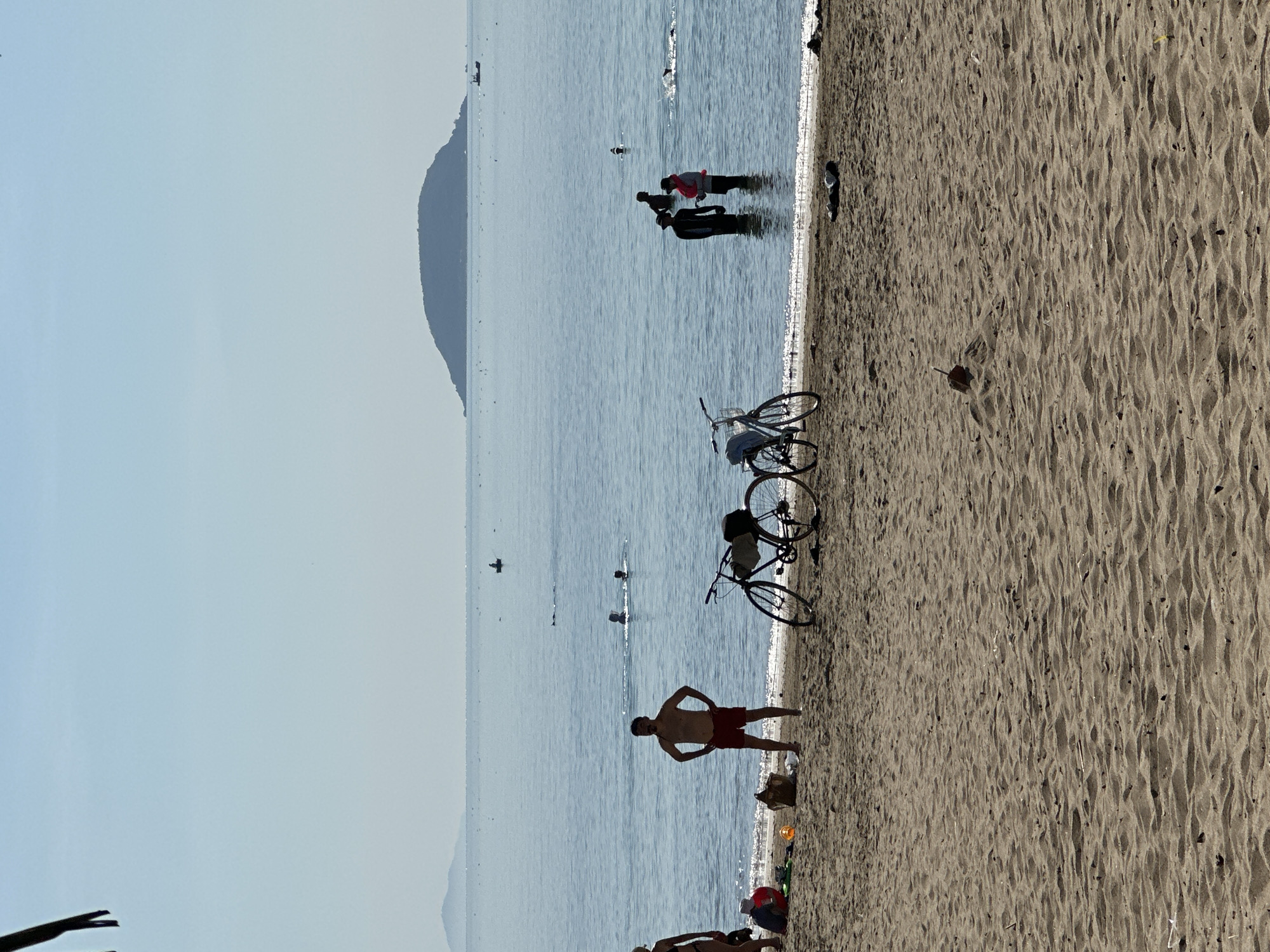 The image depicts a beach scene in Bac Nha Trang Ward, Vietnam. Two bicycles are parked on the sandy shoreline near the water's edge. A man in red shorts stands on the sand with his hands on his hips, facing the water. Further out in the water, a group of people are wading. In the background, a land mass is visible across the water. The sky is bright and slightly hazy. A few objects are scattered on the beach near the man.