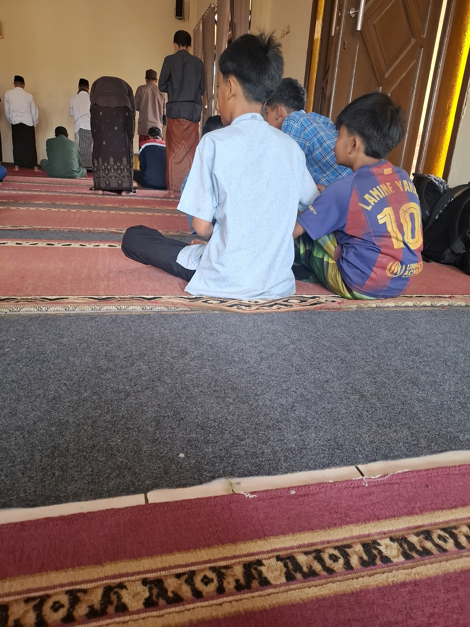 The image depicts a group of males, likely children and adults, engaged in prayer on a carpeted floor within a building. Two boys are prominently featured in the foreground, sitting on the carpet. One boy wears a light blue shirt and black pants, while the other wears a blue and red shirt with the number "10" on the back and a green and yellow patterned sarong. Several other individuals are visible in the background, some standing and others kneeling, also appearing to be in prayer. They are dressed in traditional attire, including white shirts, sarongs, and caps. The environment suggests a place of worship, possibly a mosque, with a wall, a wooden door, and curtains visible. The flooring consists of alternating red and grey carpets. The lighting appears to be natural, possibly from windows or an open doorway, casting a warm glow on the scene. The image was taken in Ciheulang Tonggoh, Indonesia.