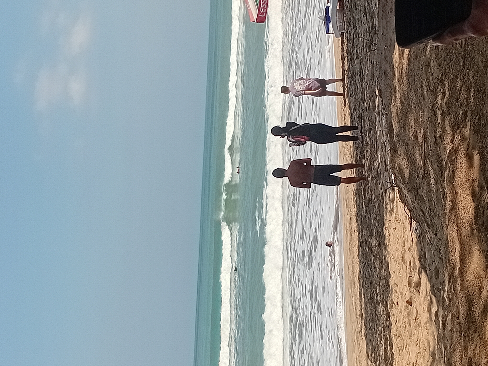 The image depicts a beach scene in Kuta, Indonesia, under a clear, pale blue sky. The ocean is a prominent feature, with multiple sets of waves rolling in, some breaking white and others a vibrant turquoise. In the distance, there are surfers in the water.
On the sandy shore, from left to right, are three individuals standing and facing the ocean. The leftmost person is a man wearing dark shorts and no shirt. Next to him is a person wearing a dark top and dark leggings, holding a dark object, possibly a phone or camera. The third person is a man wearing a patterned t-shirt and dark shorts. Further out in the surf, a child's head is visible above the water. To the right, a red and white umbrella or canopy with the word "LESS" is partially visible. The foreground is dominated by the sandy beach, with some debris and shadows cast by unseen objects, possibly trees.