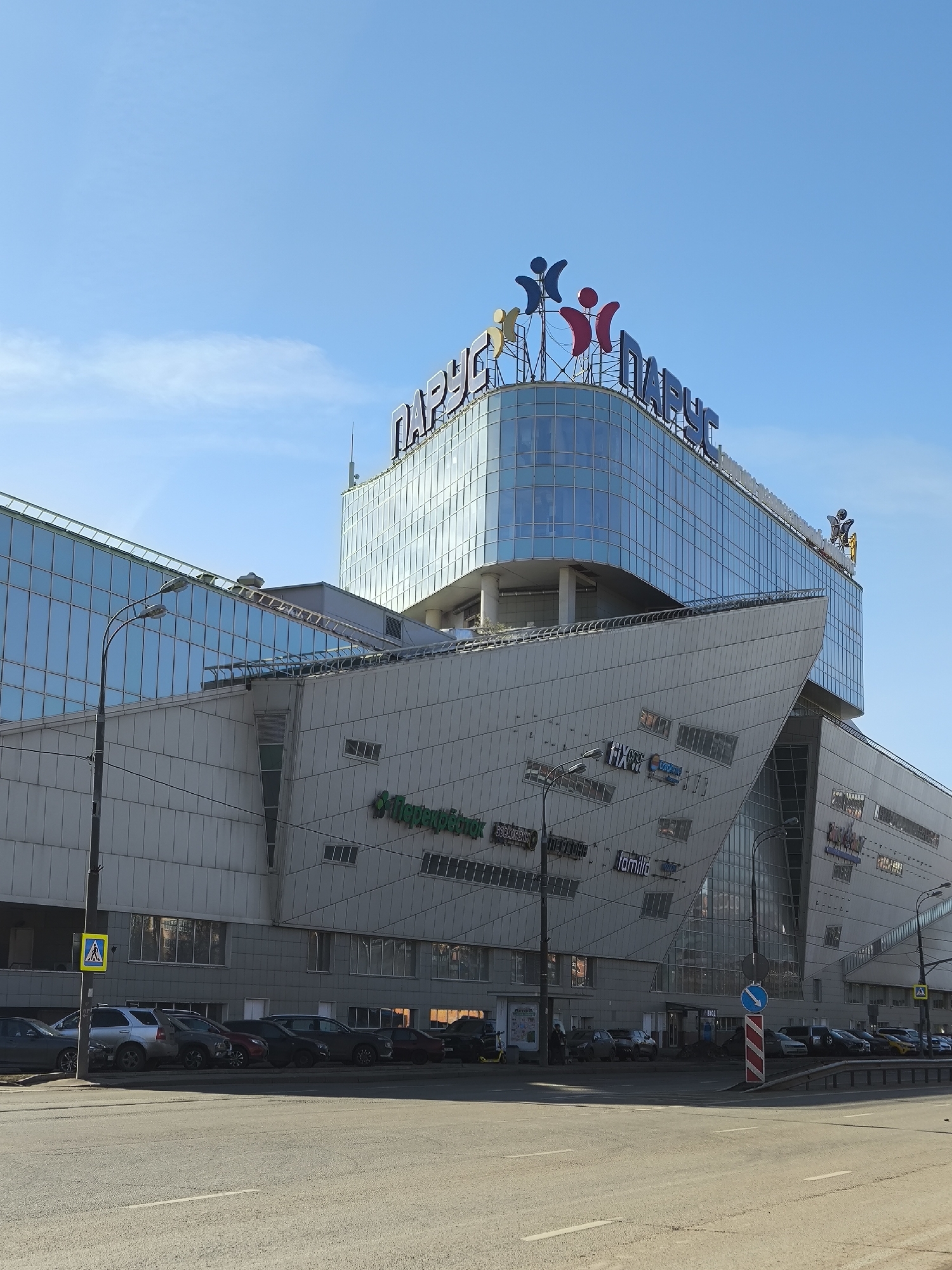 The image depicts the "Parus" shopping mall in Moscow, Russia. The building is a multi-level structure with a distinct architectural style featuring a combination of glass and concrete elements. The mall's name, "ПАРУС," is prominently displayed on the top. Various store signs are visible on the façade, including "Перекрёсток" (Perekrestok), Familia, and Fix Price. The street in front of the mall is an asphalt road, with parked cars lining the curbs. Street lamps, a pedestrian crossing sign, and a roundabout sign are present on the street. The sky is clear blue with light clouds.