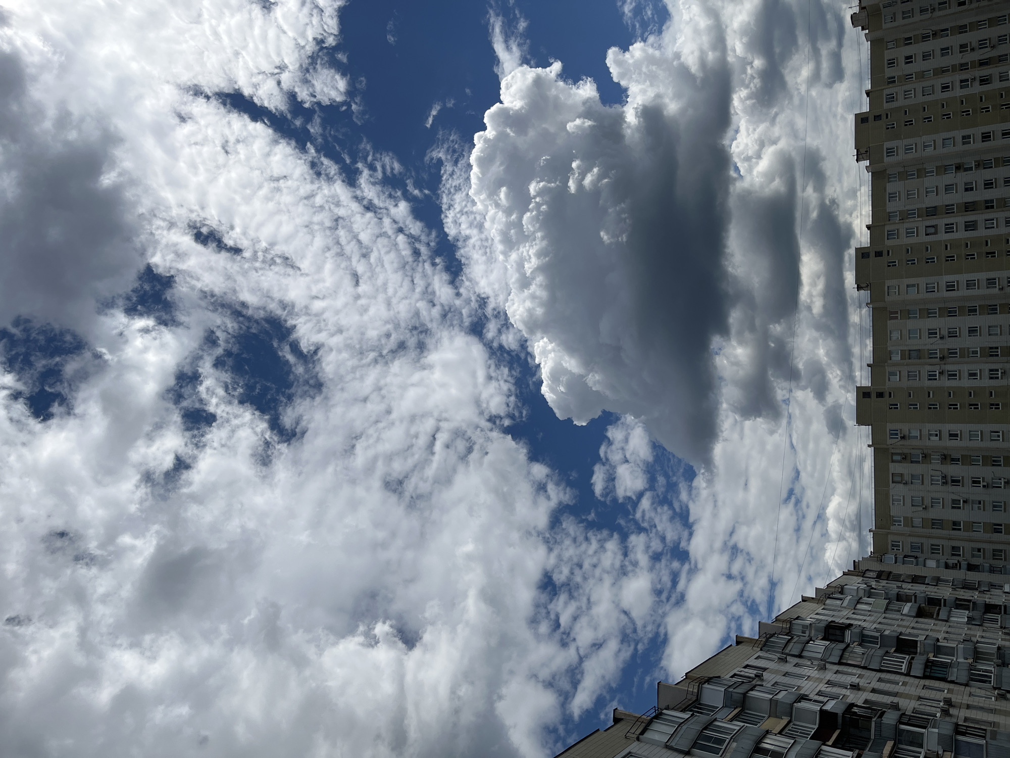 The image shows a view of apartment buildings against a cloudy blue sky. The buildings are multi-story, with many windows and balconies. The sky is filled with white and gray clouds, with patches of blue visible. There are no people or animals visible in the image. The scene appears to be an urban environment, likely in Moscow, Russia, given the architectural style of the buildings. The lighting suggests it is daytime, possibly late morning or early afternoon.