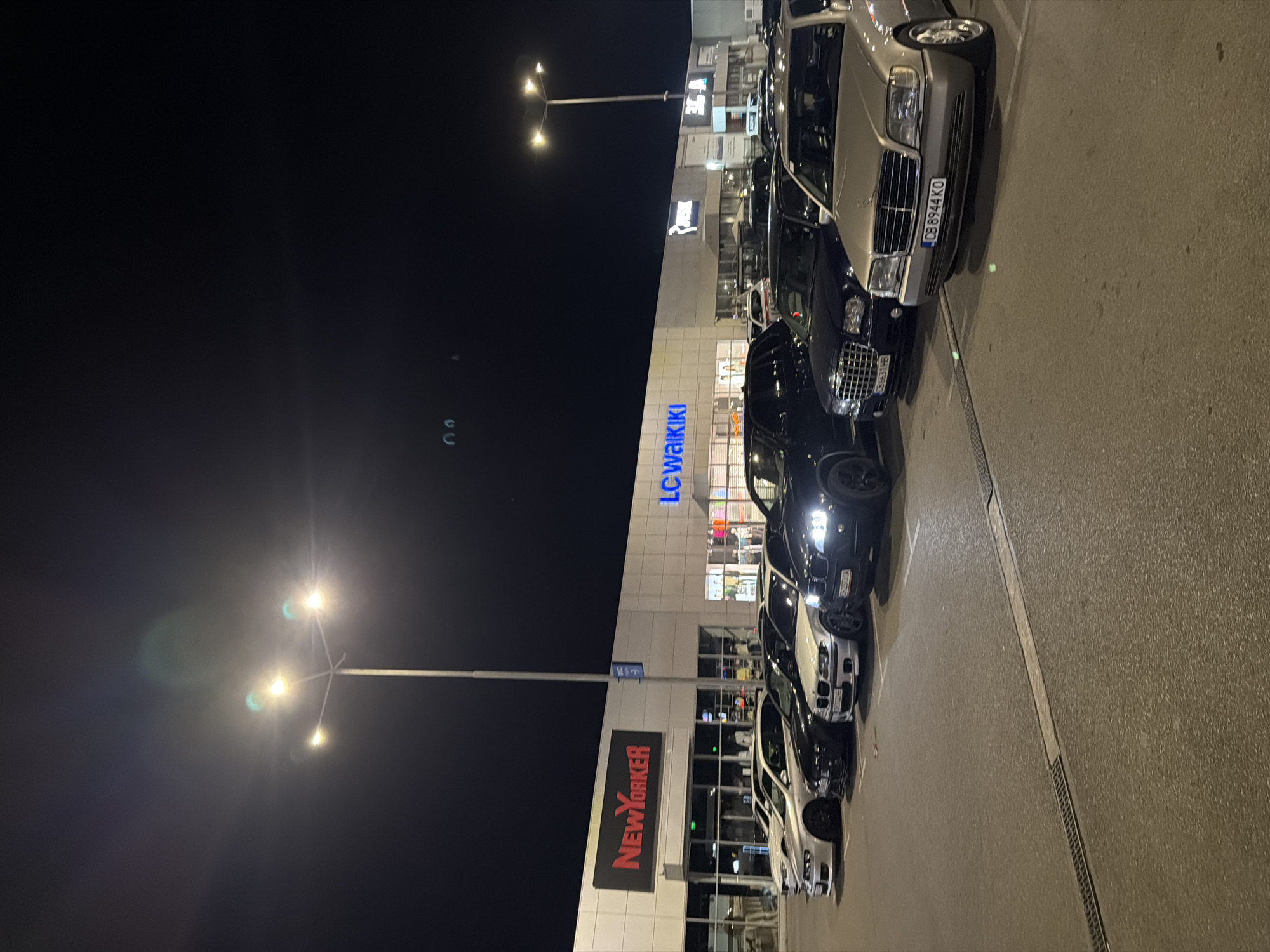 The image depicts a row of parked cars in front of a building with illuminated signs for "LC Waikiki" and "NewYorker". The license plate "CB 8944 KO" is visible on one of the cars. The location is in Sofia, Bulgaria. The time of day appears to be night, with streetlights providing illumination.