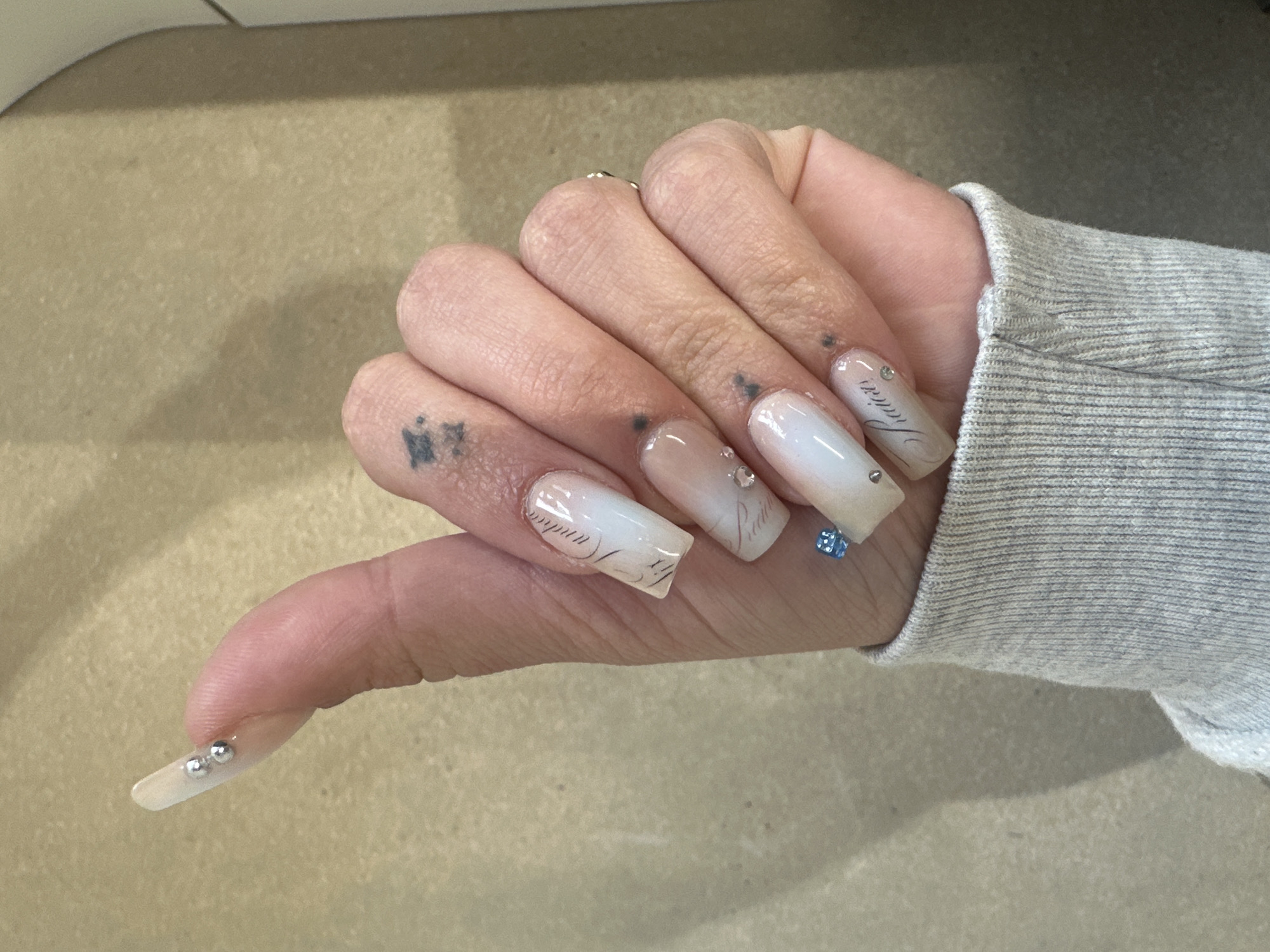 A close-up shot depicts a hand with elongated, square-shaped nails adorned with a light, milky finish, possibly a gel or acrylic application. Each nail exhibits unique embellishments; one features script-like lettering, while others have small, strategically placed rhinestones or a single, larger blue gemstone. The skin of the hand is fair, with visible fingerprints and a small, dark tattoo on the side of the index finger. The hand is positioned in front of a neutral, light-brown surface. A grey, long-sleeved garment peeks into the frame from the lower right corner.
There is no information in the image to identify a location in the city of Paris, France.