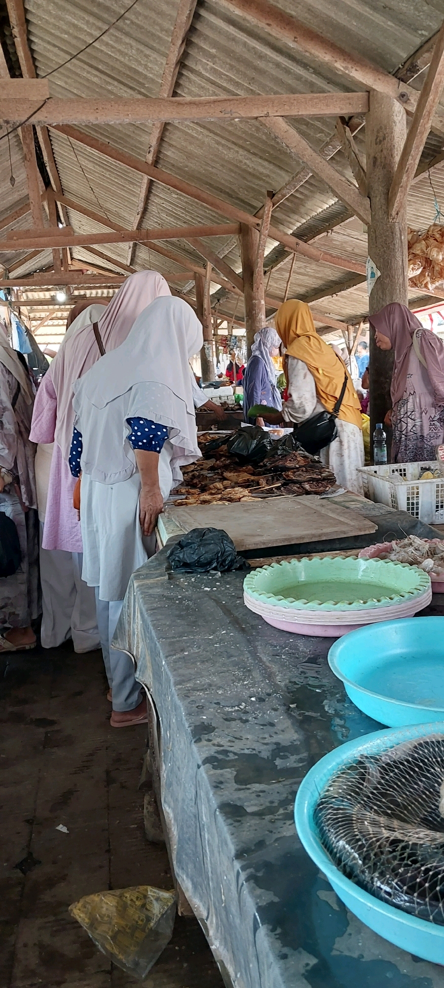 This is a full-frame, eye-level, vertical shot of a bustling fish market in Pecangaan, Indonesia. The scene is sheltered by a weathered, wooden-framed roof covered with corrugated metal sheets. In the foreground, a long, dark-colored table is covered with a plastic sheet, upon which several items are displayed. To the left, there are stacks of green and pink plastic bowls, with a blue plastic bowl on the right containing fish wrapped in a net. Behind these, a substantial amount of prepared fish, skewered and arranged on wooden sticks, is laid out. Next to the fish is a black plastic bag, and further to the right, a white plastic crate holds more seafood.
Several individuals, predominantly women, are present, all wearing hijabs. Some are actively engaged in looking at or purchasing fish. One woman in the center, wearing a light grey hijab and a white tunic, is looking down at the fish. To her left, another woman in a pink hijab is partially visible. To the right of the central figure, a woman in a yellow hijab is interacting with a customer, holding a green leafy vegetable. Another woman in a purple hijab is visible further back, and a woman in a mauve hijab is seen on the far right. The floor appears to be dirt or concrete, with some debris and water visible. The overall atmosphere suggests a typical, active marketplace environment.