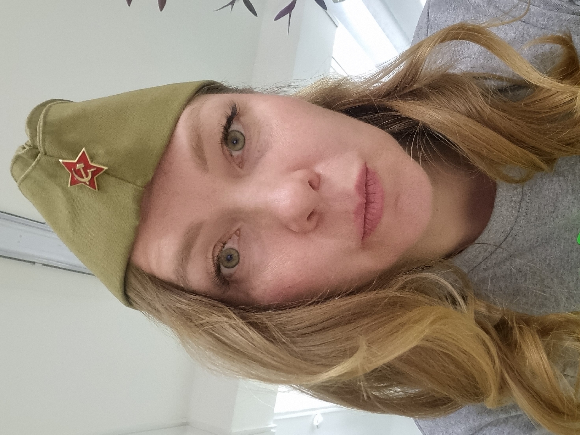 A woman with blonde hair is wearing an olive-green garrison cap with a red Soviet star pinned to the front. The cap is angled on her head. She is wearing a gray t-shirt. The background is a white wall with foliage and white blinds. The scene is not identifiable as the city of Kemul, Russia, based solely on the given image.