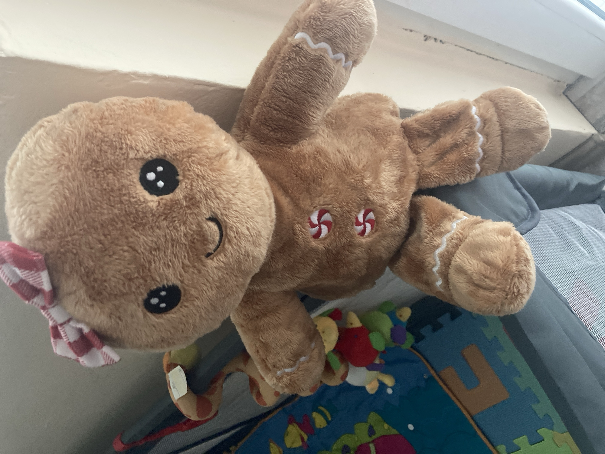 The image shows a close-up of a plush gingerbread man toy with a red and white checkered bow on its head. The toy has embroidered eyes and a smile, and two red and white swirl buttons on its chest. White icing-like stitching decorates its arms and legs. In the background, a section of a multi-colored play mat with interlocking puzzle pieces is visible, along with a baby mobile with hanging toys. The scene appears to be indoors, possibly in a nursery or playroom. There is no information in the image to suggest a location in Burgas, Bulgaria.