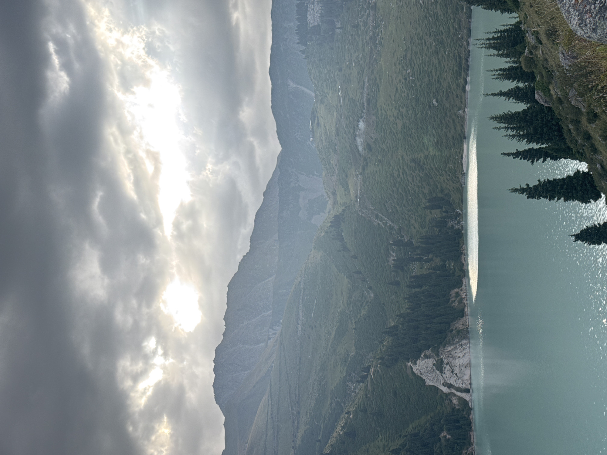 The image depicts a scenic mountain lake in Almaty, Kazakhstan, likely Big Almaty Lake, due to its distinctive turquoise waters and surrounding mountainous terrain. The sky is overcast with dramatic clouds, with a bright shaft of sunlight piercing through, illuminating a portion of the lake. Lush green vegetation covers the rolling hills and steep mountain slopes. A jagged, rocky outcrop juts out from the left side of the lake. Several conifer trees are scattered across the landscape, with a denser cluster along the lakeshore in the foreground and on the right side of the image. The water is calm, with gentle ripples reflecting the light. There are no people or animals visible in the image. The overall atmosphere is one of natural beauty and tranquility.