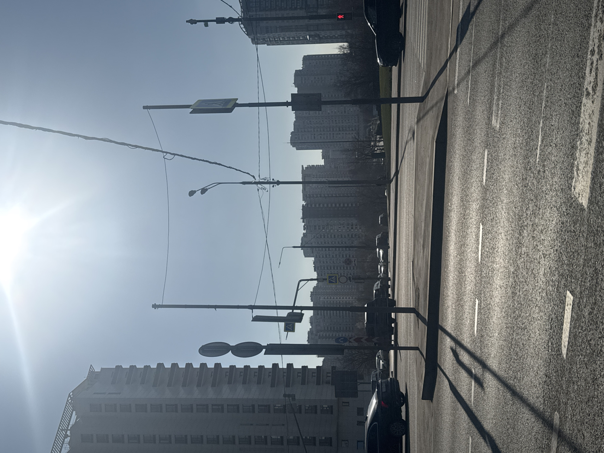 The image shows a street scene in Moscow, Russia. The road is asphalt with white lane markings and a median strip. There are several cars parked and moving along the side of the road. Tall apartment buildings form the backdrop, receding into the hazy distance. A series of streetlights and signs are positioned along the roadside. The sky is bright and washed out by the sun, causing glare in the lower left of the image. There are no people visible in the photo. The scene appears to be taken during the daytime. The buildings appear to be residential.