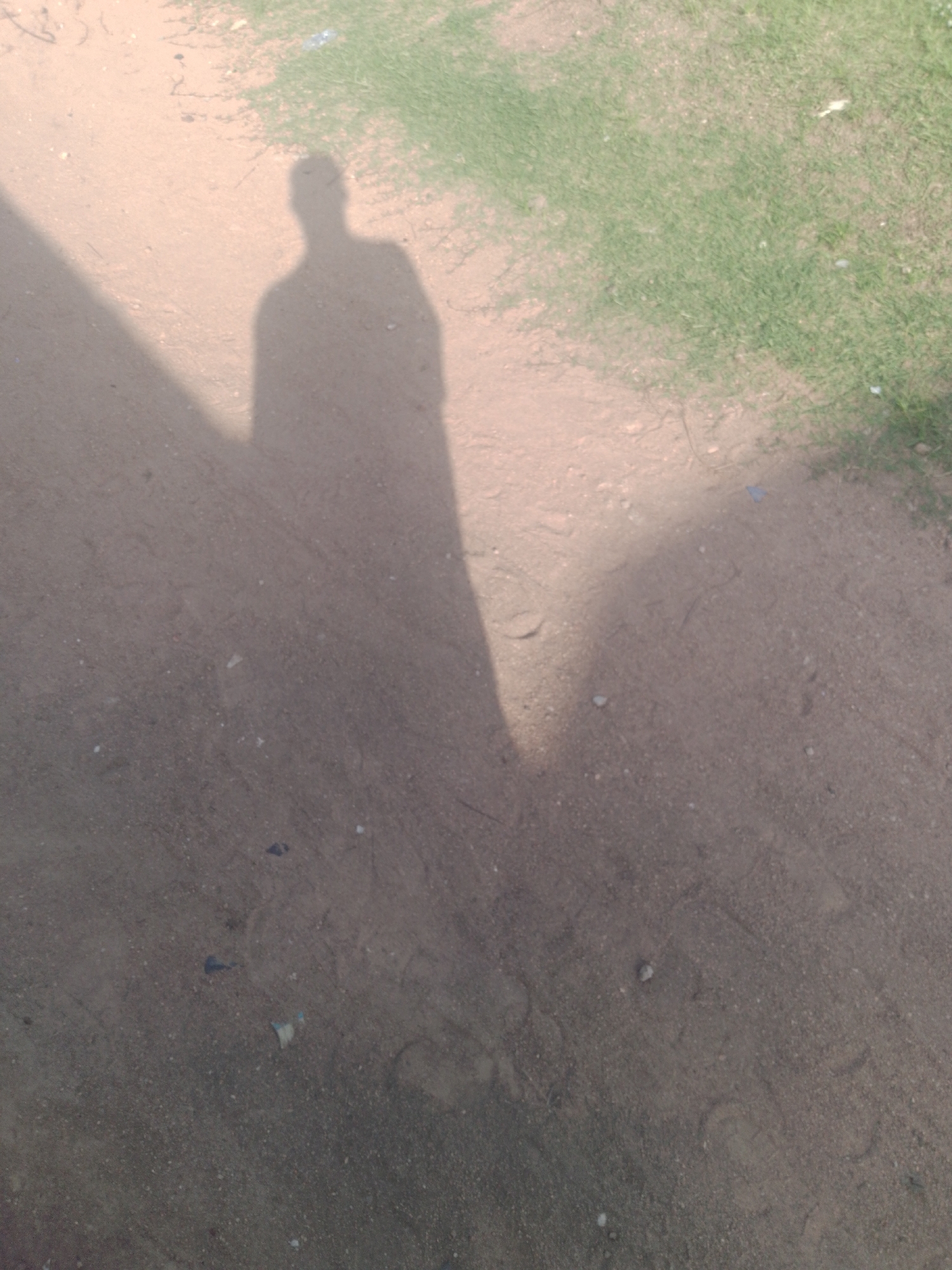 In Jalingo, Nigeria, a shadow of a person is cast on a dirt road beside a patch of grass. The shadow is long and extends across the majority of the frame, suggesting the sun is low in the sky. The dirt road is a light brown color and appears to be somewhat uneven, with small pebbles and debris scattered across its surface. There are faint impressions in the dirt that could be footprints. The grass on the right side of the image is a vibrant green and appears to be well-maintained. The overall lighting suggests it is daytime, likely late afternoon or early morning.