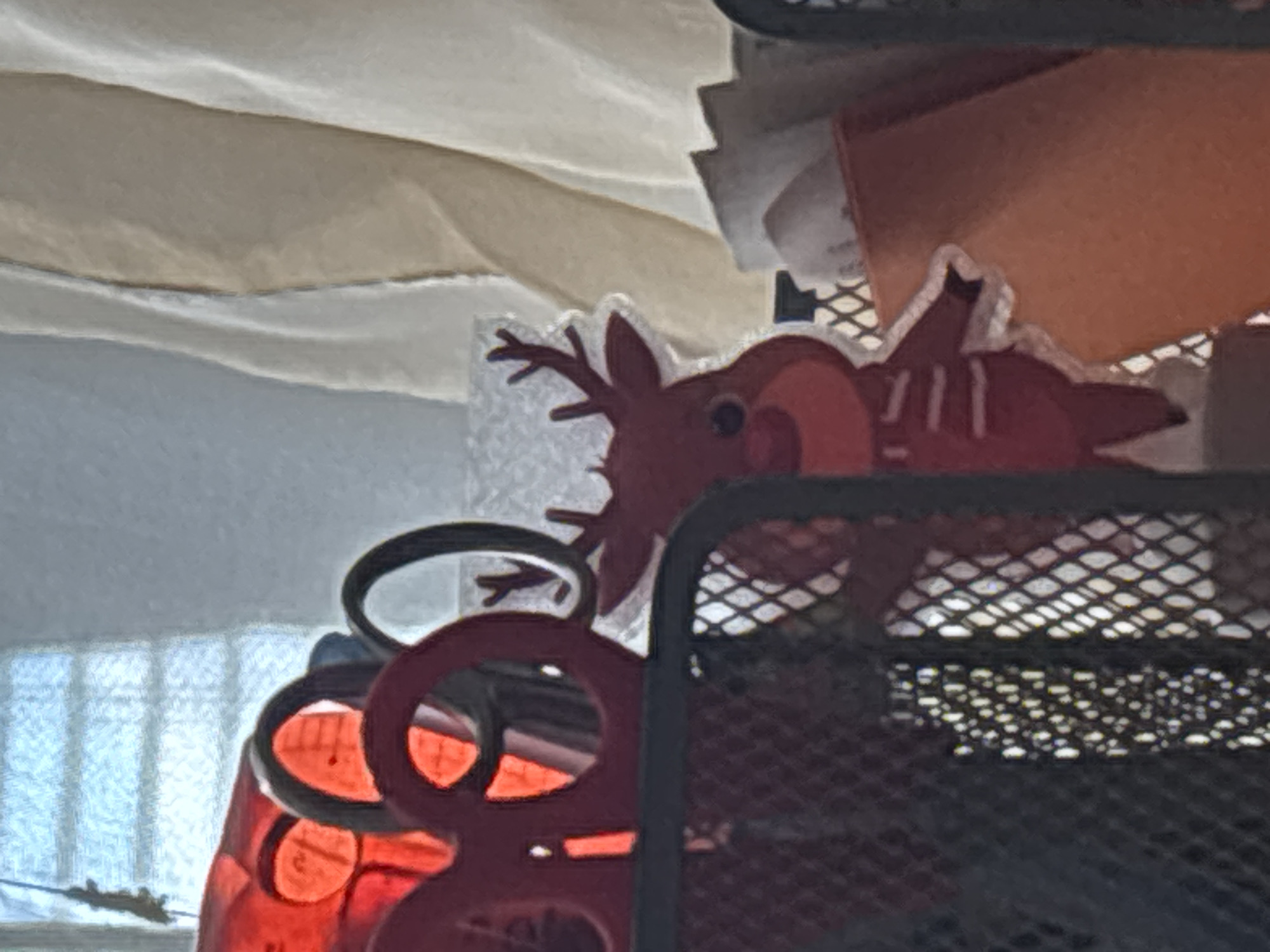 An indoor scene, observed in the city of Burgas, Bulgaria, displays a collection of desk or craft supplies. In the foreground left, a black mesh organizer partially obscures its contents. Within this organizer, several pairs of red-handled scissors are prominently visible, with their circular finger loops and elongated blades arranged vertically. Behind the scissors and partially to the right, a red reindeer-shaped cut-out decoration is visible. This reindeer features antlers, a small dark eye, and several thin white horizontal lines across its body. To the immediate right of the reindeer, a light orange or reddish-brown container holds several white papers or cards. Further to the right, a cream-colored fabric, likely a curtain or drape, hangs vertically, exhibiting soft folds. In the background, a window with horizontal elements, possibly blinds or windowpanes, allows diffuse light into the scene. A thin, dark, vertical object hangs near the window's left side. The overall lighting suggests daylight. No people are present.