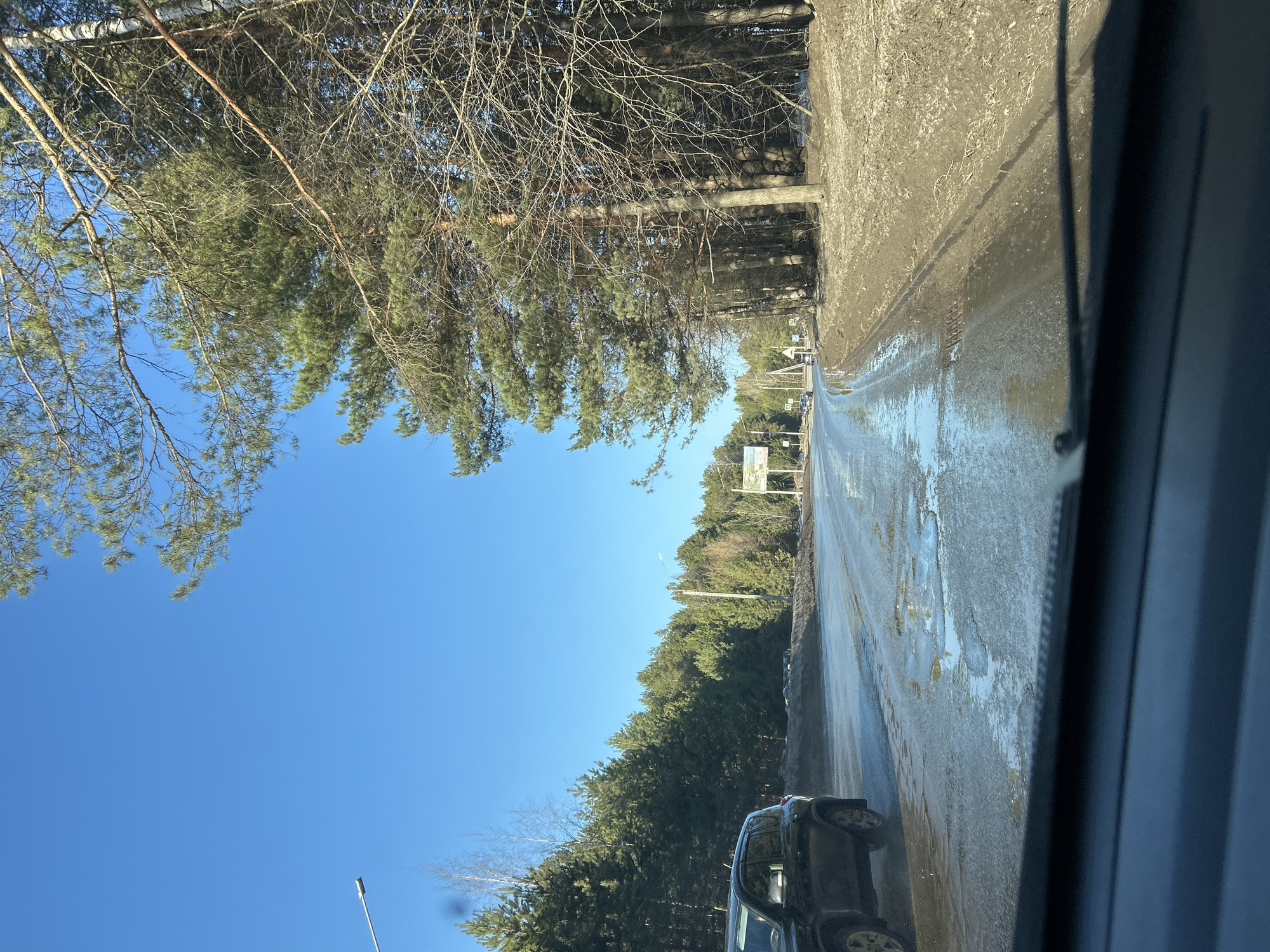 The image, likely taken from within a vehicle, shows a partially flooded road in what appears to be a wooded area. Patches of ice and water are visible on the road surface. A black SUV is partially visible on the lower right side of the frame. Trees line both sides of the road, with bare branches and some evergreen foliage. A clear blue sky is visible above the trees. The perspective is slightly tilted. Based on the city's location, this image could have been taken in Chaikovsky, Russia. Other details, like street signs are indiscernible, prohibiting any other description.