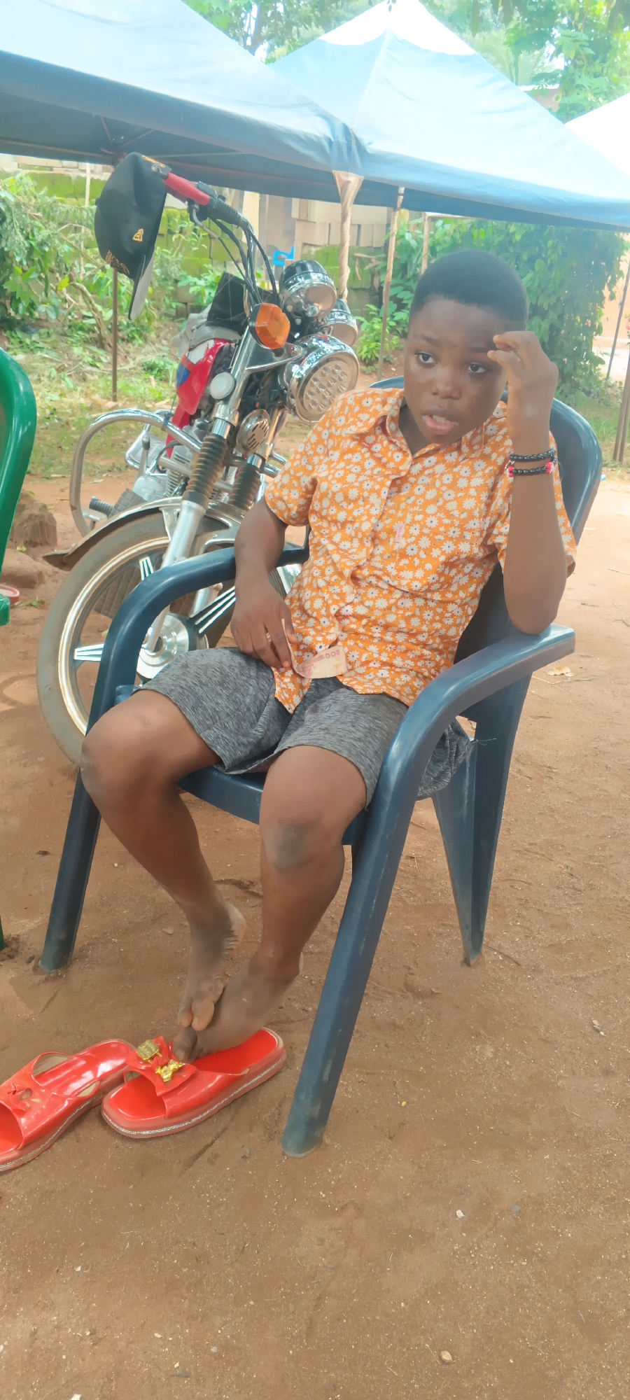A young boy with dark skin and short dark hair is seated in a blue plastic chair in an outdoor setting in Eziudo, Nigeria. He is wearing an orange short-sleeved collared shirt with a white floral pattern and grey shorts. He has black and pink/reddish bracelets on his right wrist. His left hand holds a green 500 Nigerian Naira note, partially visible. His bare feet are dusty and rest on the reddish-brown dirt ground, near a pair of bright red slide sandals with gold embellishments. There's a dark scuff mark on his right knee.
Behind the boy, a silver and red motorcycle with chrome details is parked. A black baseball cap hangs from its handlebar. The scene is under a light-colored canopy tent, providing shade. In the background, green foliage and some concrete block structures are visible. The boy's expression appears thoughtful or pensive, with one hand touching his forehead. The overall impression is a casual daytime scene in a possibly rural or semi-urban outdoor area.