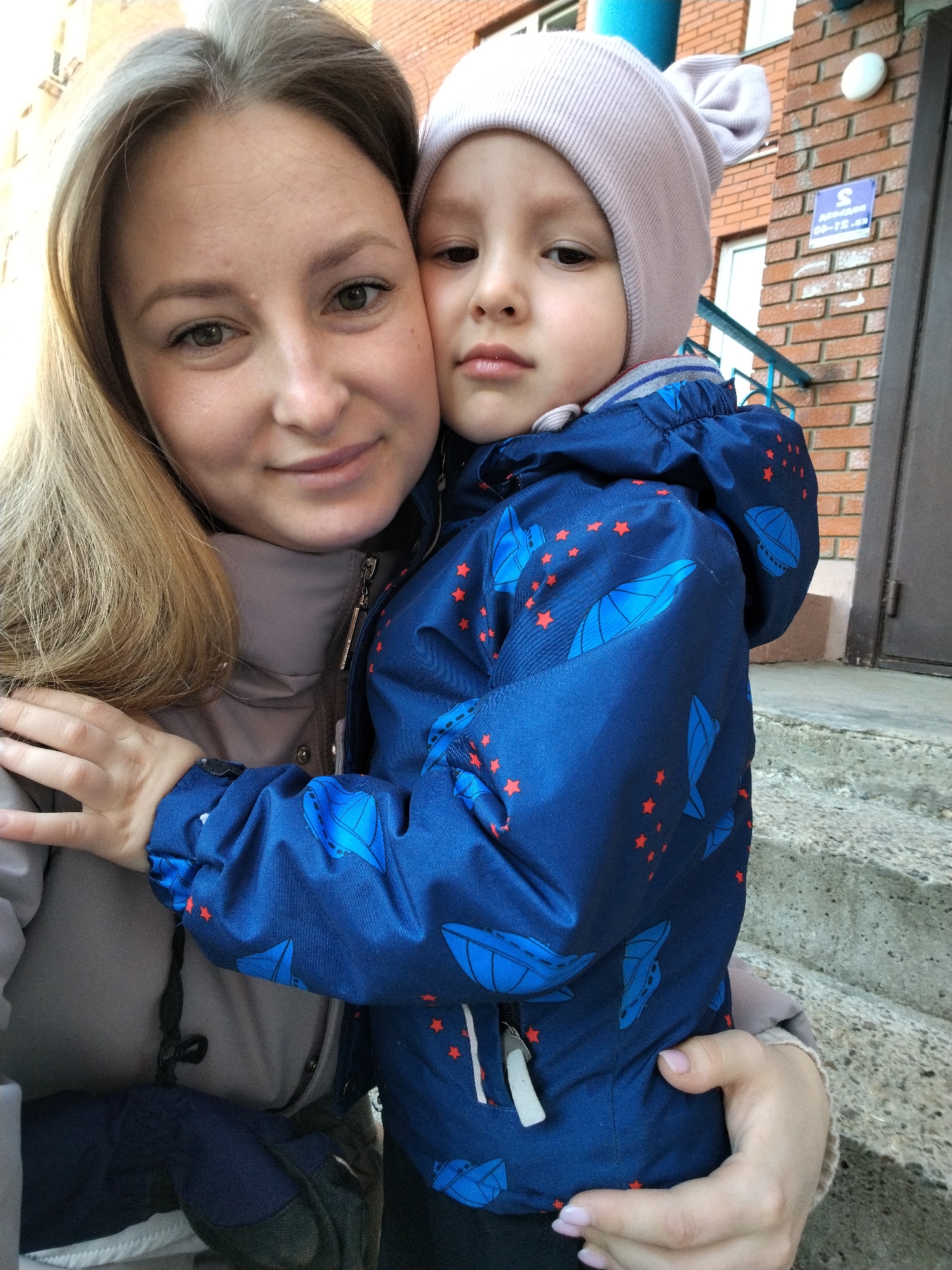 The image depicts a woman and a child in front of a brick building. The woman is fair-skinned with light brown hair and is wearing a gray jacket. She has her arm around the child. The child is wearing a light pink hat with ear-like protrusions, and a blue jacket decorated with a pattern of blue spaceships and red stars. The building has a brick exterior, a white window frame, and a dark gray door. The location is in the city of Chaikovsky, Russia.