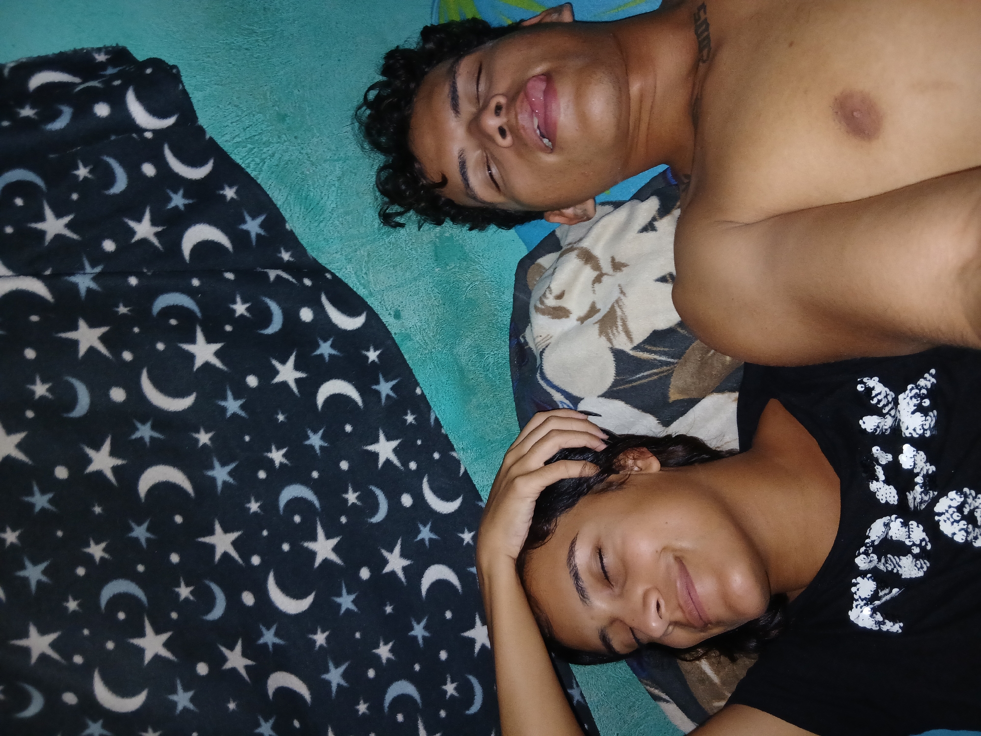 Two individuals lie side-by-side on a bed in what appears to be an indoor setting. The first individual, a shirtless male, lies on his back with his mouth slightly open. He has curly hair. The second individual, a female, lies with her eyes closed. She is wearing a dark t-shirt. Her left arm is bent, with her hand touching her hair. The bedding beneath them consists of several layers, including a teal/aqua blue bedspread, a patterned pillow or blanket with beige and dark brown floral designs, and a dark blanket with a pattern of white and light blue stars and crescent moons. The location is described as Altagracia, Venezuela.