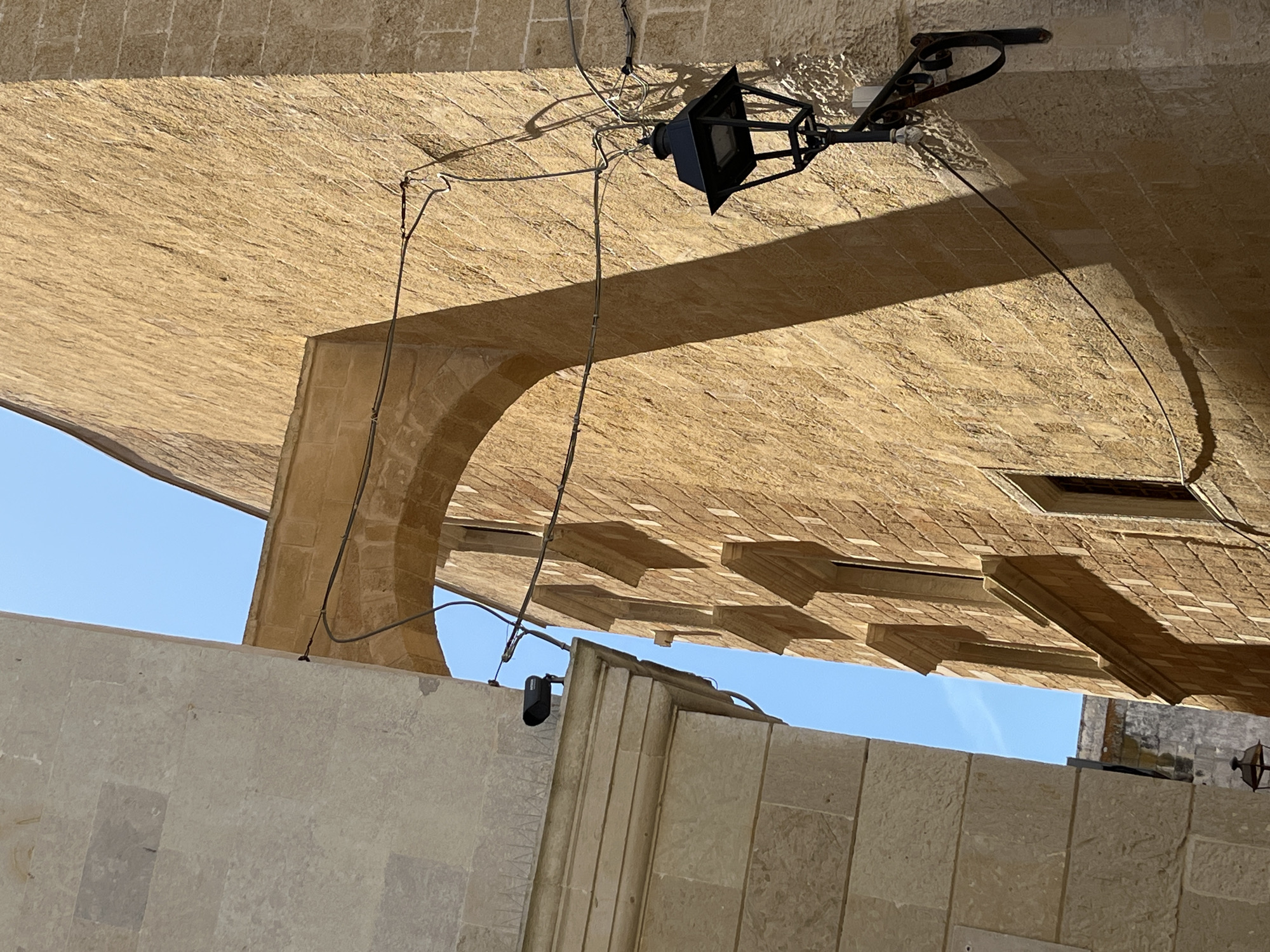 The image shows an outdoor scene in Otranto, Italy, looking up at an architectural overhang. The structure is constructed of light-colored stone, possibly limestone, common in the region. There is a wrought-iron lamp fixture attached to the underside of the overhang with exposed wiring and electrical boxes. The sky is visible in sections between architectural details. The presence of the stone structures and the lamp suggest an old and possibly historic area.