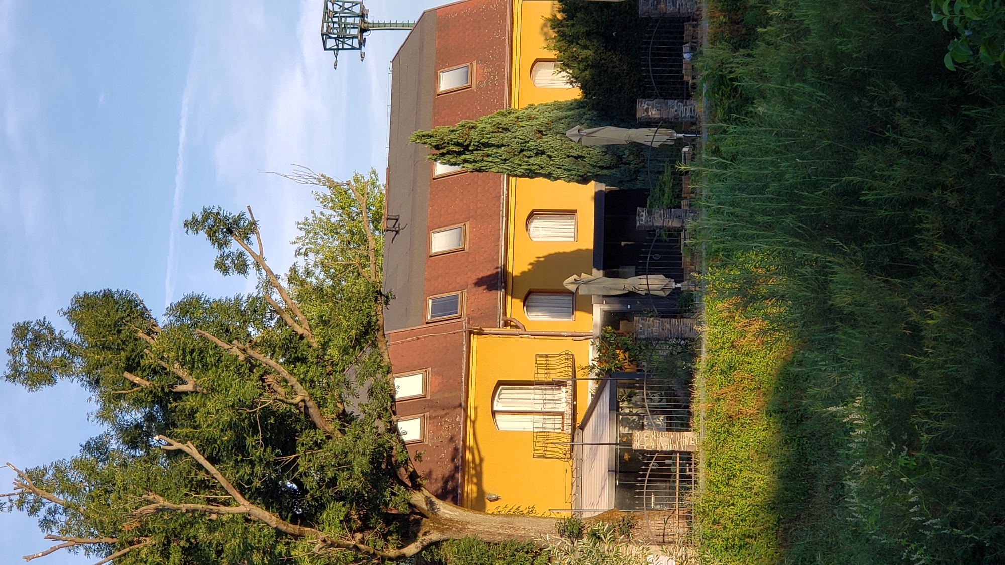 The image shows a two-story building with a yellow facade and a reddish-brown tiled roof. The roof features several dormer windows. The building appears to be a restaurant or hotel, with outdoor seating areas visible behind a black wrought-iron fence. There are umbrellas set up on the patio. A large tree with lush green foliage is prominent on the left side of the building, and a tall, slender evergreen tree stands to the right. The area in front of the building is covered in dense green vegetation, suggesting it might be near a body of water or parkland. In the background, a metal structure resembling a transmission tower is visible against a blue sky with scattered white clouds. The lighting suggests it is late afternoon or early evening, with sunlight casting long shadows. This scene is characteristic of the area along the Torysa River in Prešov, Slovakia, specifically near the city's sports complex which often features such recreational and dining facilities. There are no people visible in the image.
