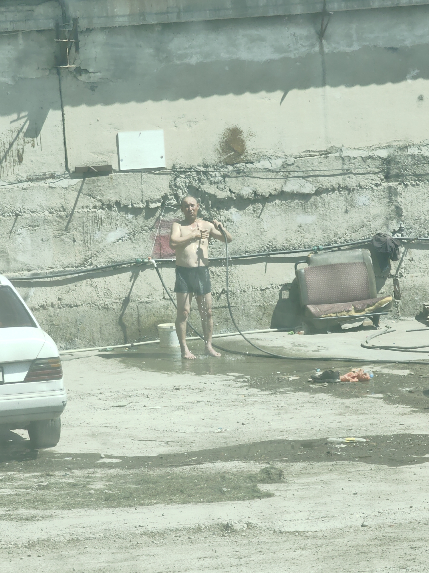 A bald man wearing black shorts is standing barefoot in a puddle of water and being sprayed by a hose, suggesting he is showering outdoors. He appears to be in an urban setting, possibly in Shymkent, Kazakhstan, as indicated by the context of the request. The environment is somewhat dilapidated, with a rough concrete wall in the background, some pipes and wires running along it, and a dilapidated car seat to the right. A white car is partially visible on the left side of the image. The ground is uneven and appears to be a mix of dirt and concrete, with some debris scattered around, including a white bucket and some plastic bags. The lighting suggests it is daytime. The man is holding the hose with his right hand and appears to be washing his chest or face with his left. Water is splashing around him.