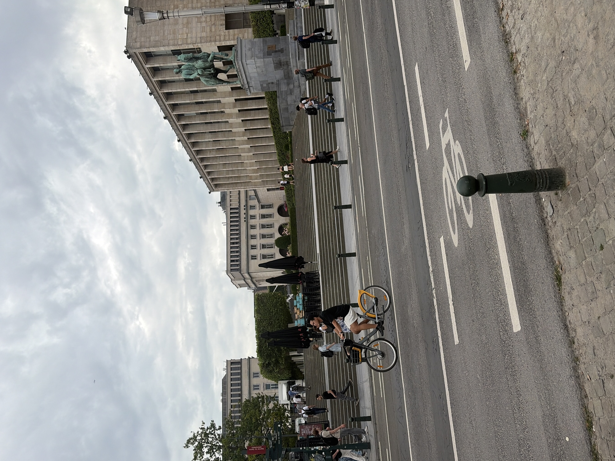 The image shows a street scene in Brussels, Belgium, likely in the area of Mont des Arts or Place Royale, given the architecture and the presence of the horse statue which resembles the equestrian statue of King Leopold II. In the foreground, a paved road with a bicycle lane is visible, marked with white lines and a bicycle symbol. A dark green bollard stands on the edge of the sidewalk. A person is riding a yellow and grey bicycle with a basket on the front along the street. In the background, there is a large, imposing stone building with a row of columns, and a grand staircase leading up to it. Several people are seen walking up and down the stairs and on the sidewalk. The sky is overcast. The overall impression is that of an urban public space on a cloudy day.